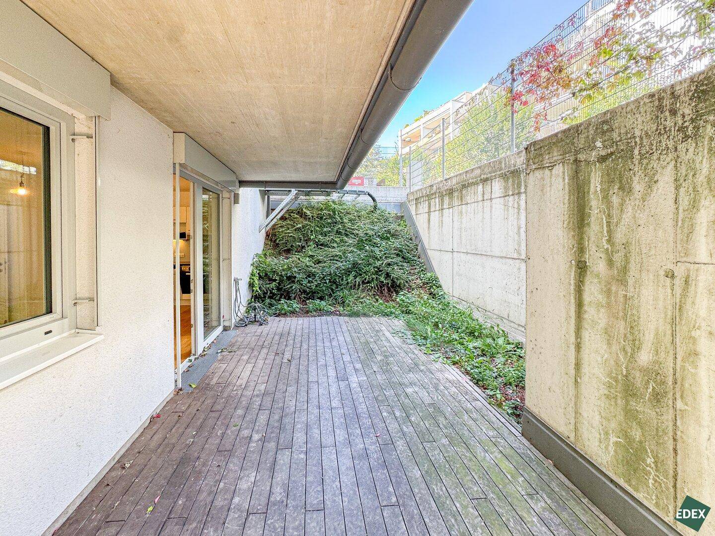 Eine Holzterrasse im Freien, umgeben von Betonwänden und üppigem Grün, bietet einen privaten Rückzugsort.