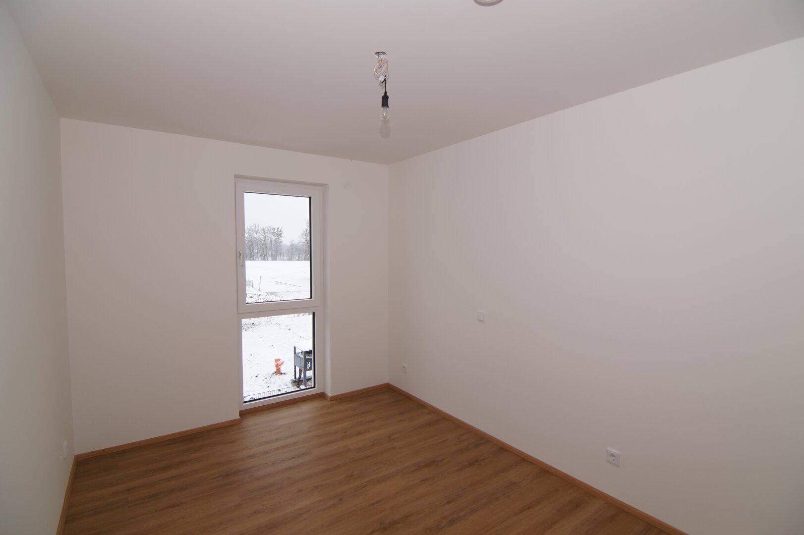 Leeres Zimmer mit Laminatboden und großem Fenster, das viel Tageslicht hereinlässt.