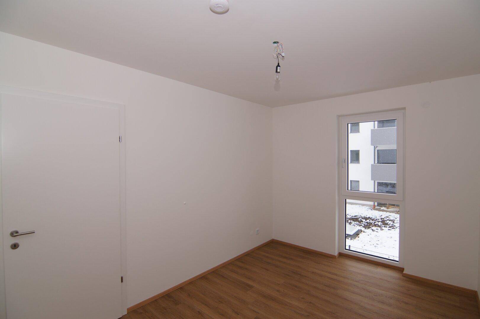 Helles Zimmer mit Laminatboden und Fenster, das einen Blick nach draußen ermöglicht.