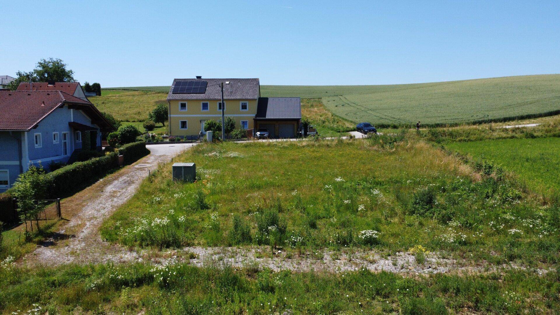 Luftaufnahme eines unbebauten Grundstücks mit einem Wohnhaus und Feldern im Hintergrund unter blauem Himmel.