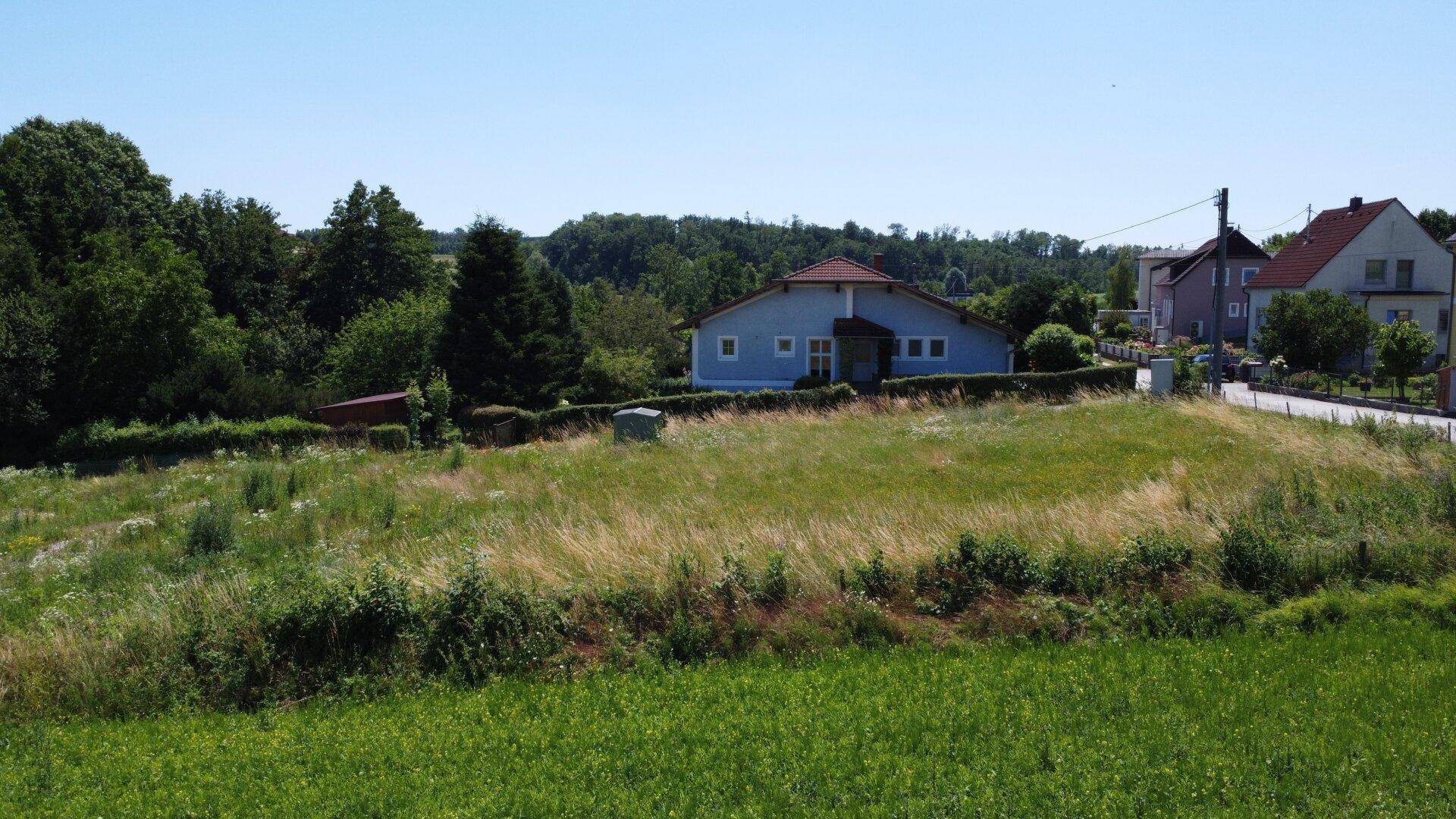 Luftaufnahme eines unbebauten Grundstücks mit einem blauen Wohnhaus und weiteren Gebäuden in der Ferne.