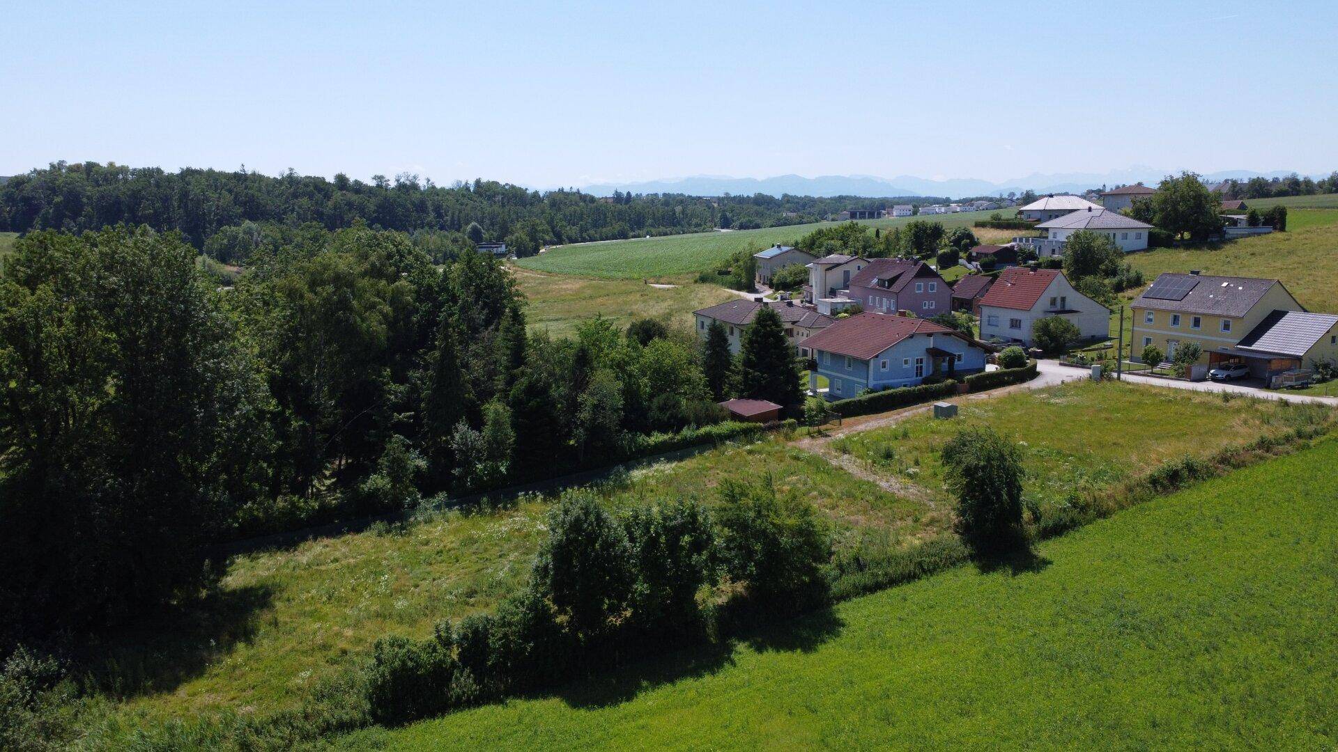 Weitläufige Luftaufnahme einer ländlichen Siedlung mit Wohnhäusern, Feldern und bewaldeten Hügeln im Hintergrund.