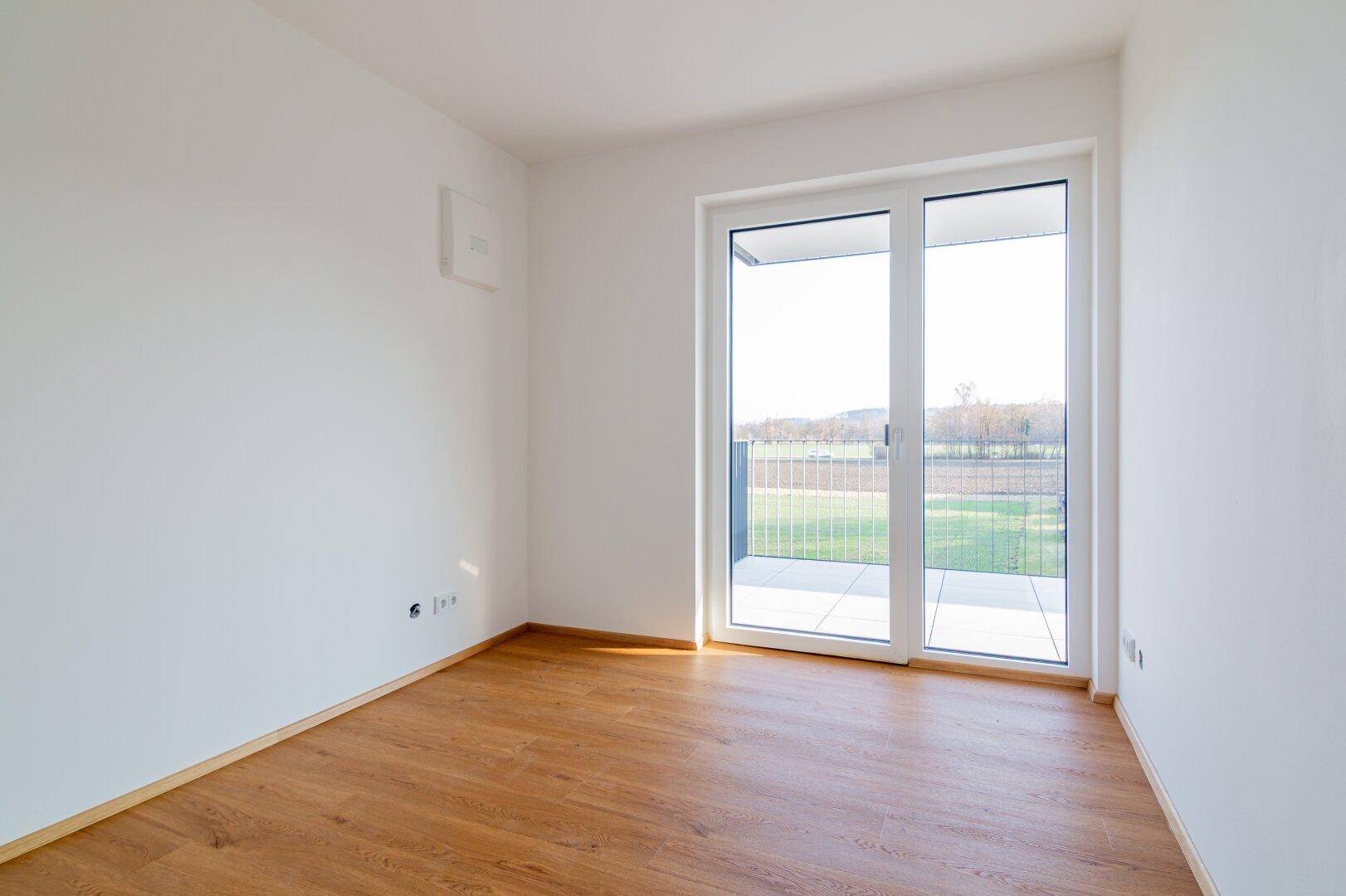 Heller Raum mit Holzboden und großer Balkontür, die einen weiten Blick in die Natur bietet.