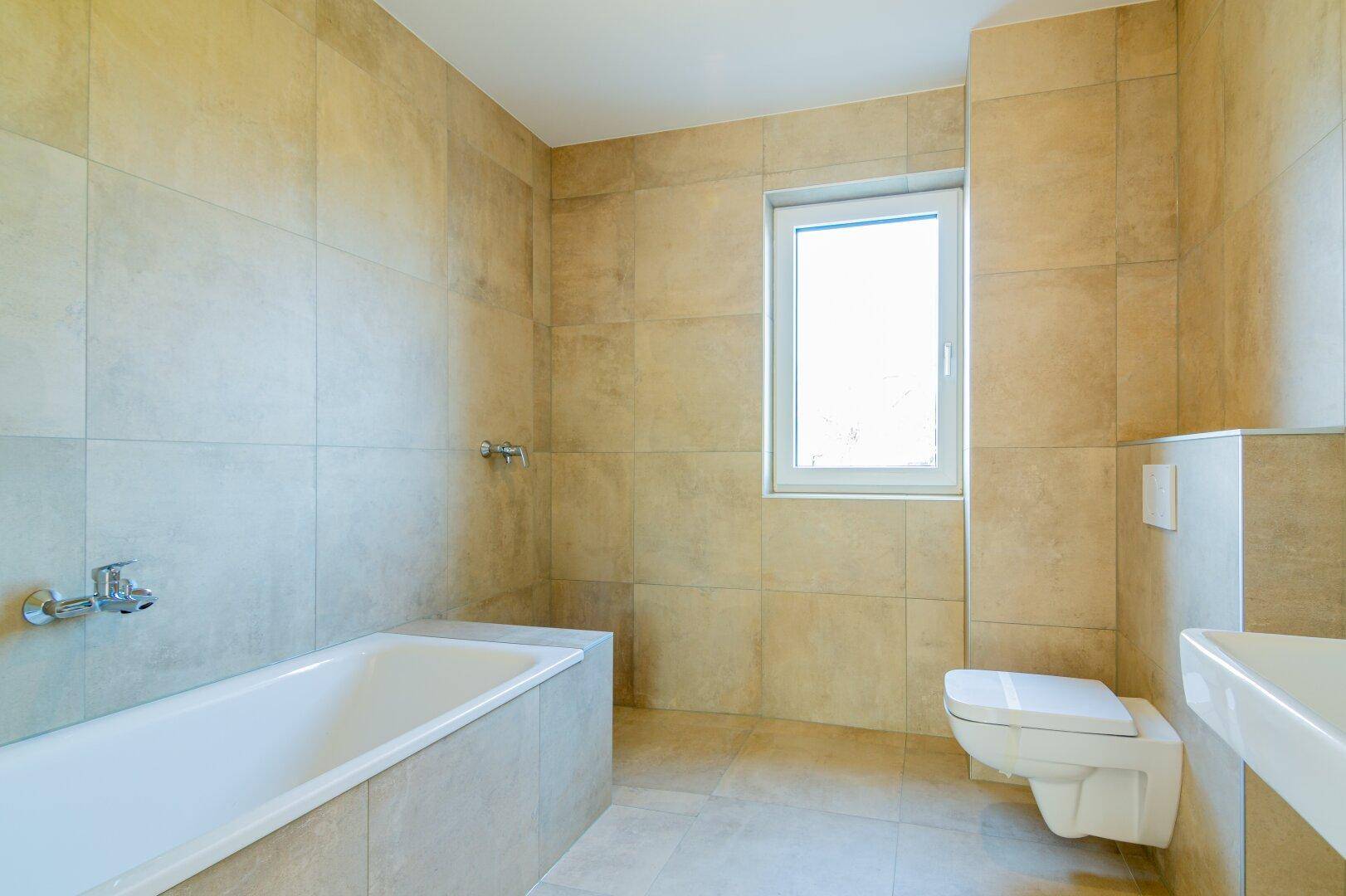 Modernes Badezimmer mit beigefarbenen Fliesen, Badewanne, wandhängendem WC und Fenster.