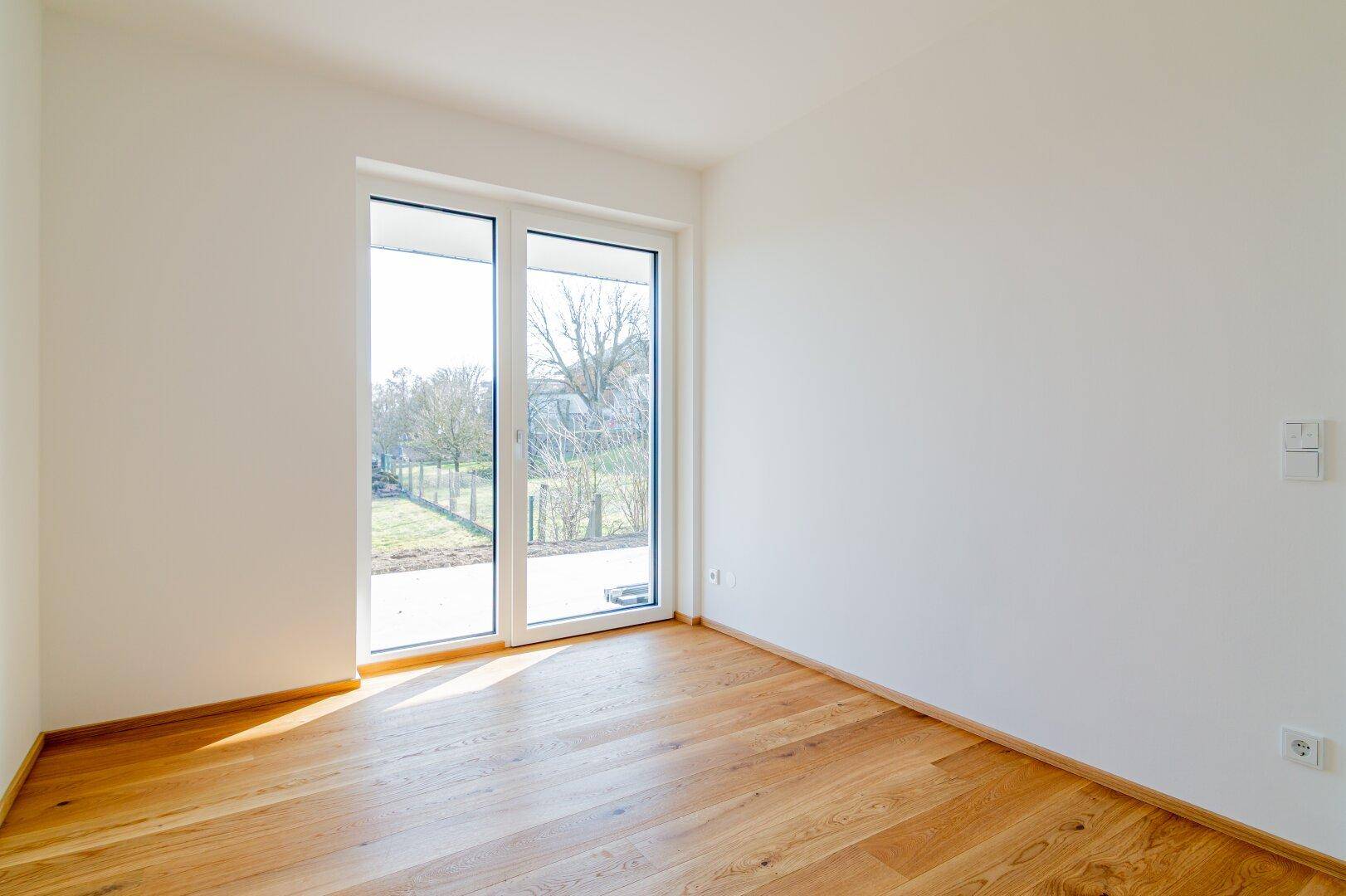 Lichtdurchflutetes Zimmer mit Holzboden und großem Fenster mit Zugang zum Außenbereich.
