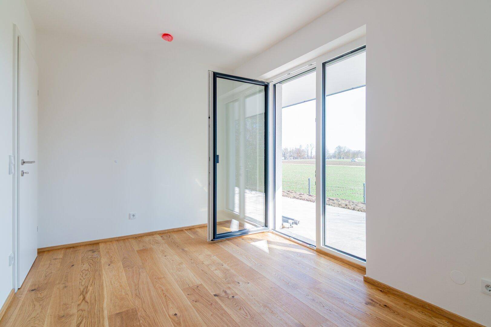 Helles Zimmer mit Holzboden und Zugang zu einem Außenbereich mit Blick ins Grüne.