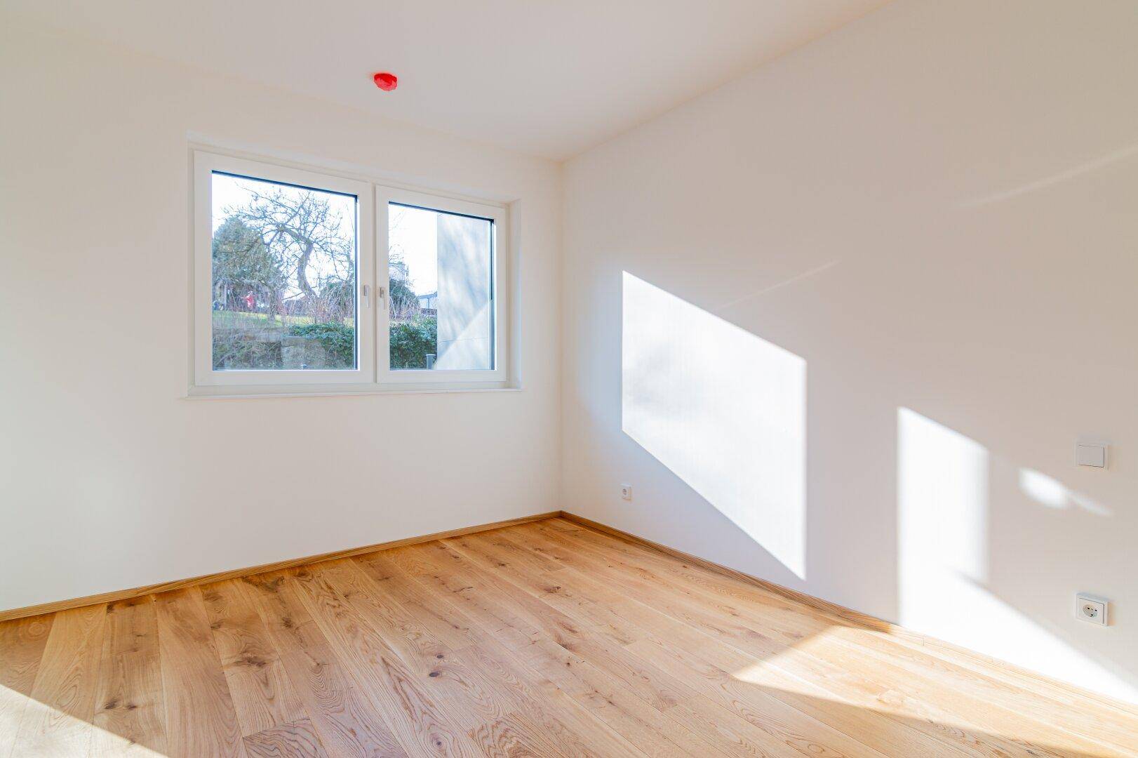 Helles Zimmer mit Holzboden und Fenster, das einen Blick ins Grüne bietet.