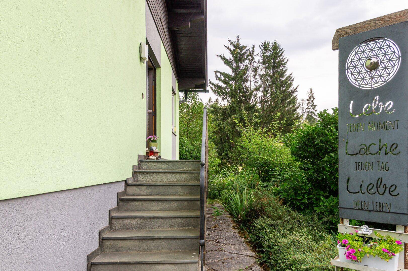 Außentreppe zum Hauseingang mit einer dekorativen Tafel und umgebendem Grün der Natur.