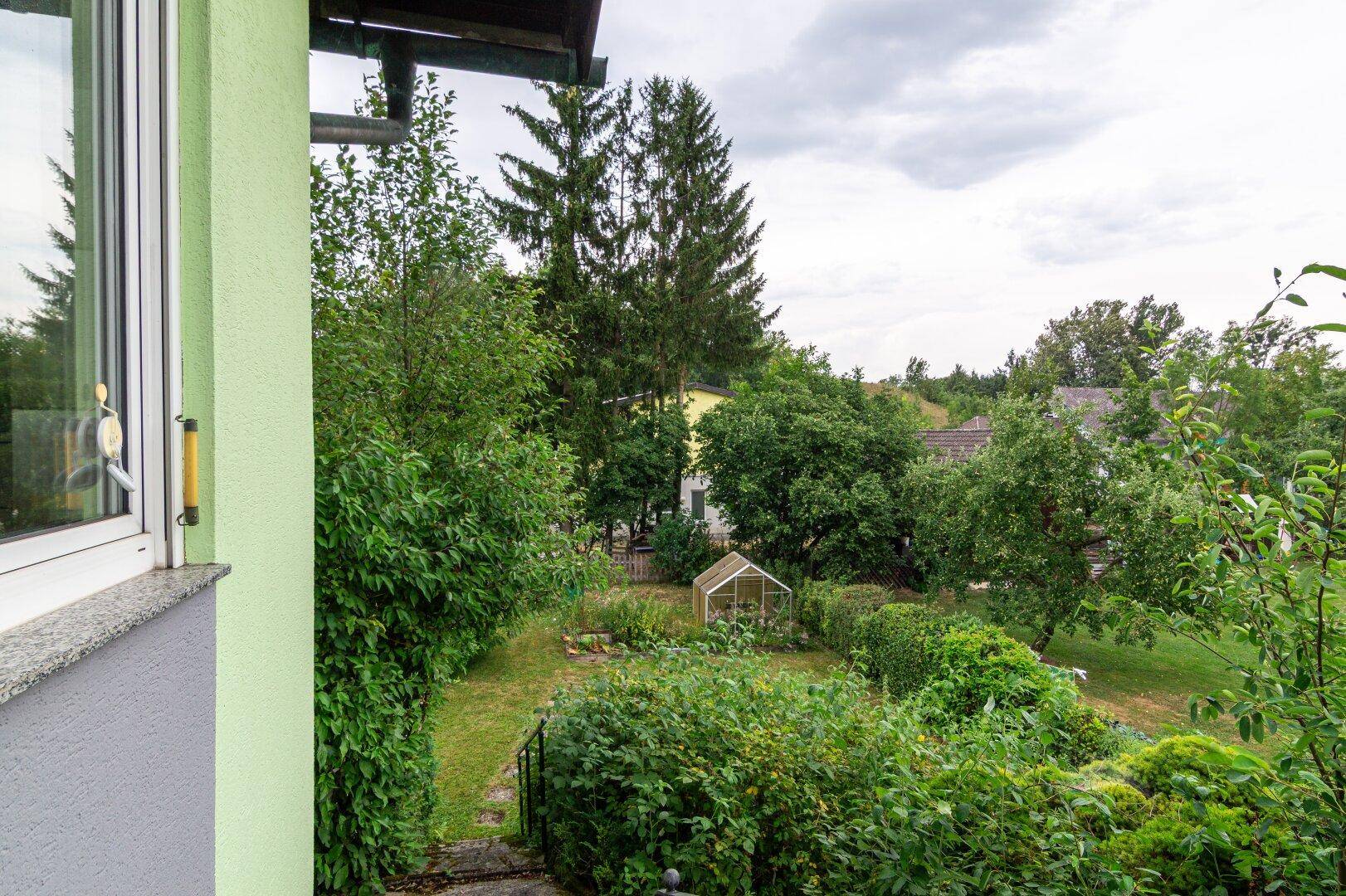 Blick aus dem Fenster auf einen gepflegten Garten mit Bäumen und einem kleinen Gewächshaus.