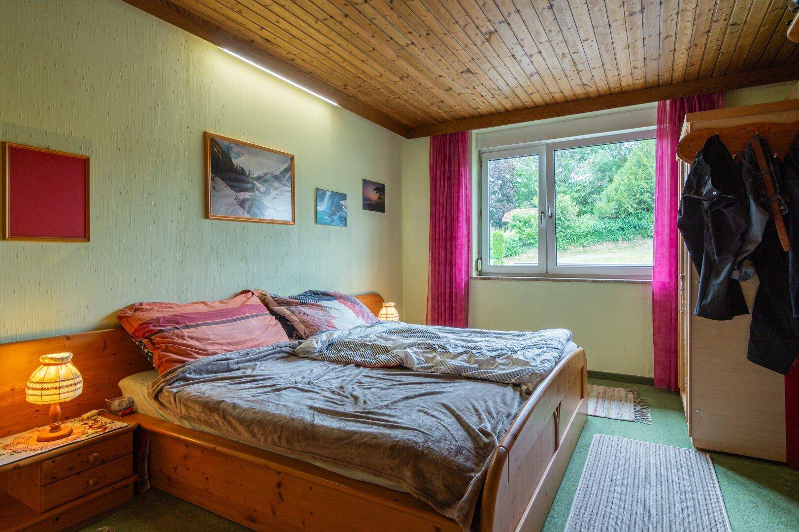 Schlafzimmer mit Holzbett, Holzdecke und Blick ins Grüne durch das große Fenster.