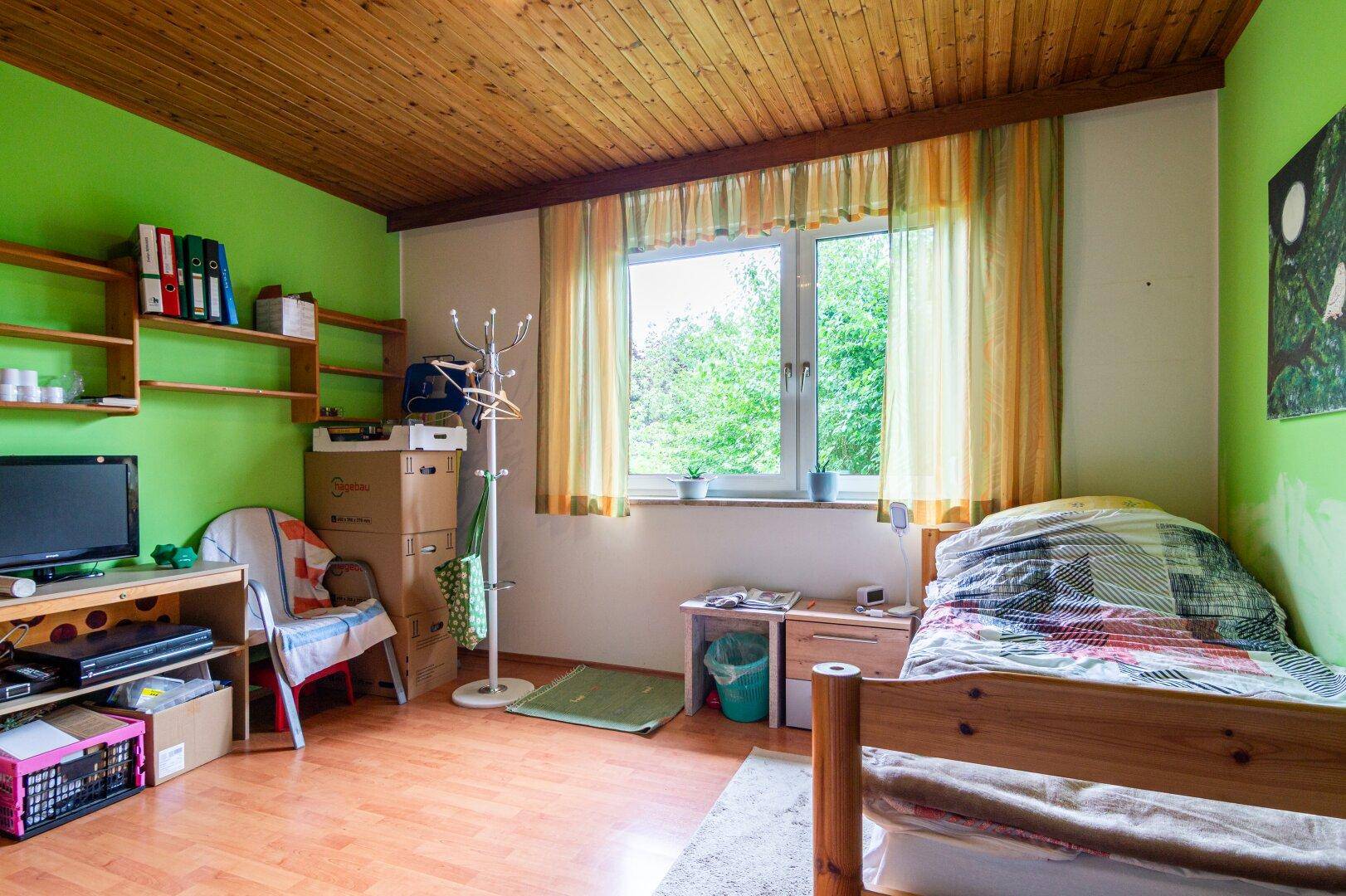 Helles Schlafzimmer mit Holzdecke, Laminatboden und Blick auf die grüne Umgebung.