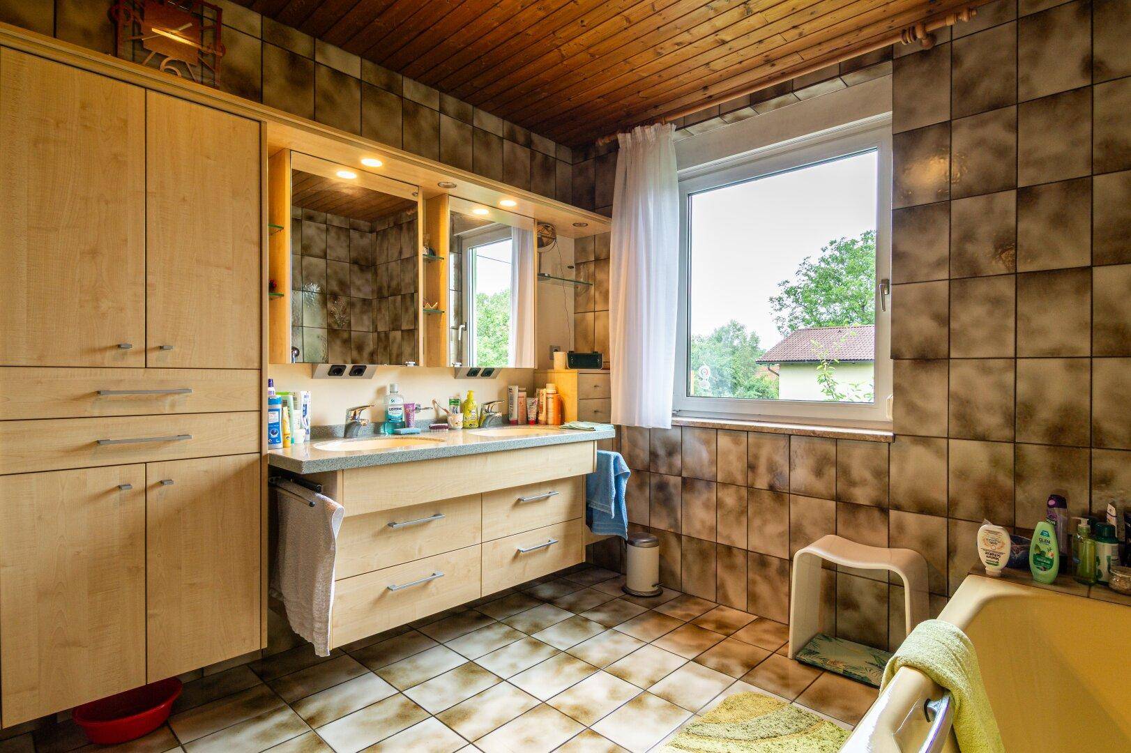 Badezimmer mit braunen Fliesen, Doppelwaschbecken und Badewanne, ausgestattet mit einem Fenster.