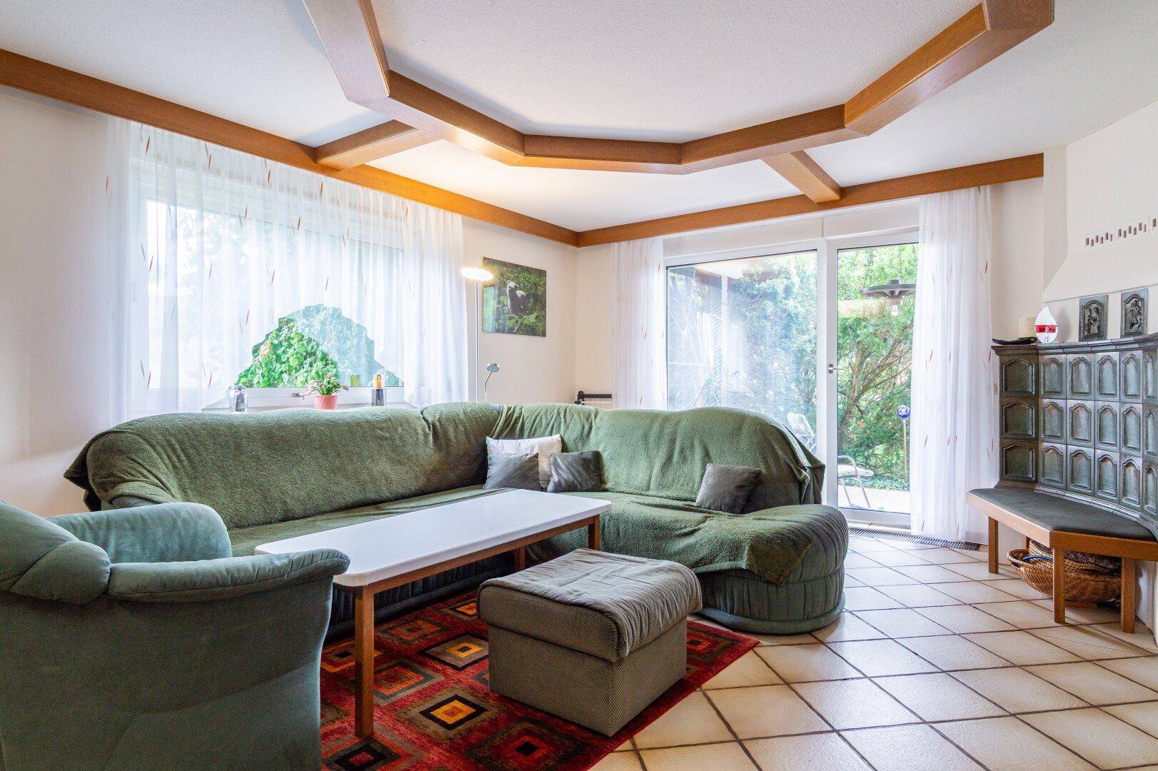 Gemütliches Wohnzimmer mit grünem Sofa, Kachelofen und Zugang zur Terrasse im Freien.