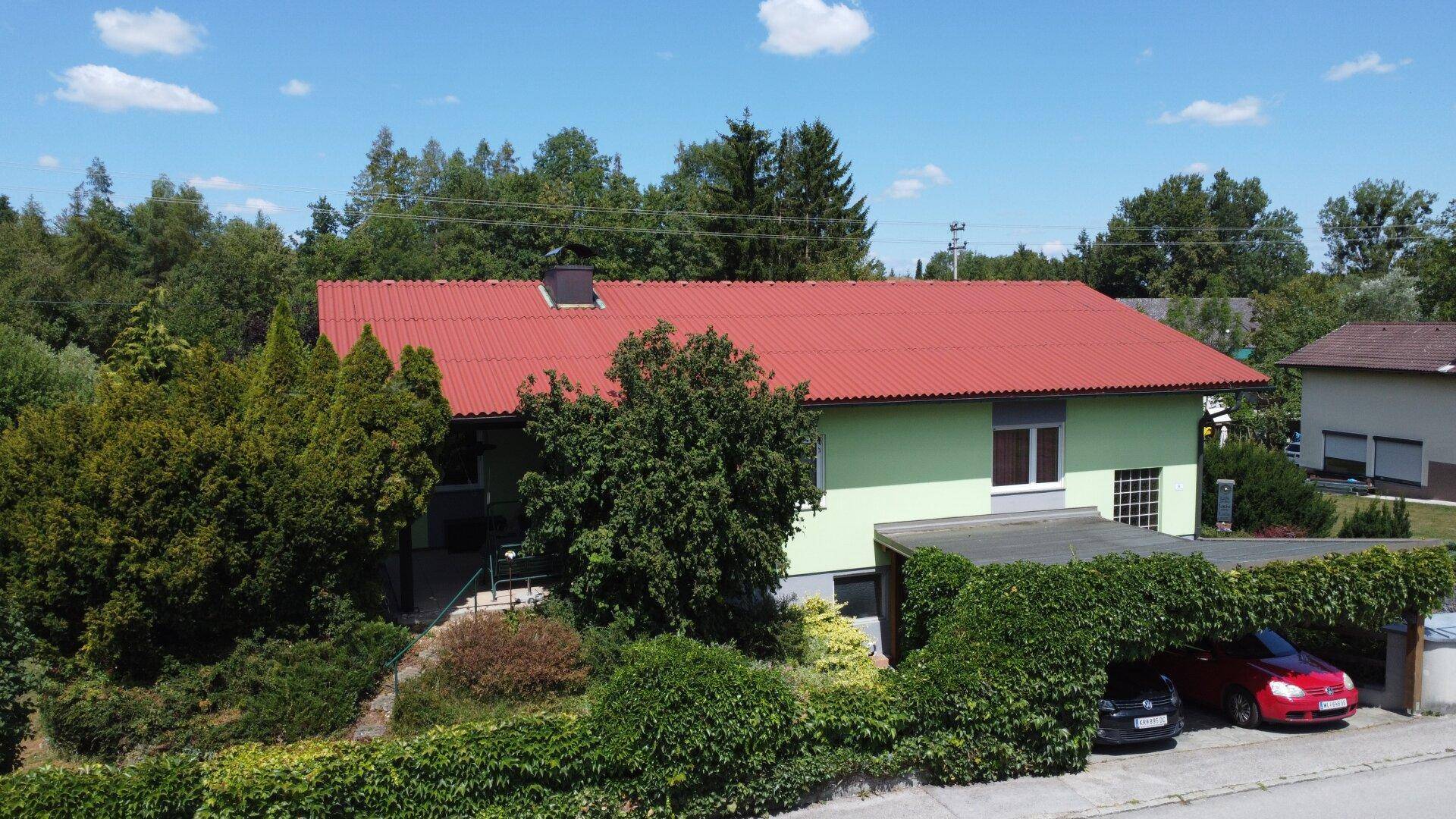 Haus mit rotem Dach und hellgrüner Fassade, umgeben von Bäumen und Sträuchern, mit Carport.