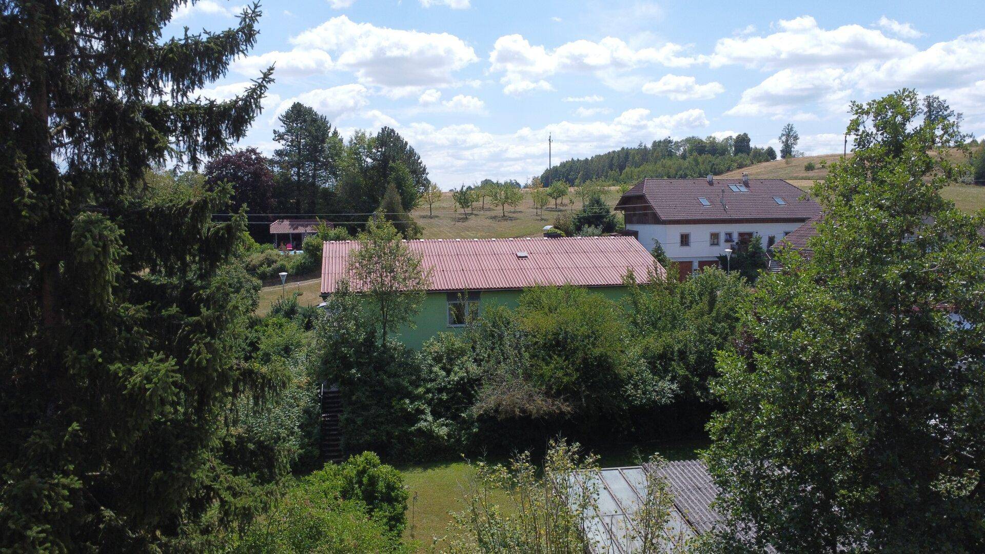 Draufsicht auf das Haus mit rotem Dach, umgeben von üppiger Natur und Feldern unter blauem Himmel.