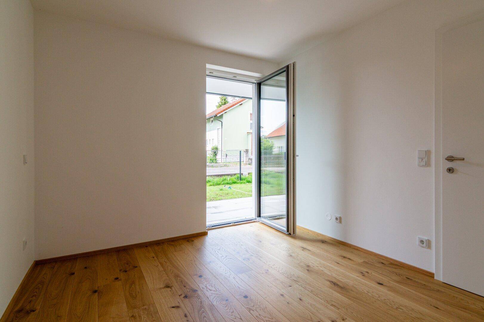 Helles Zimmer mit Holzboden und offener Glastür, die den Blick ins Grüne freigibt.