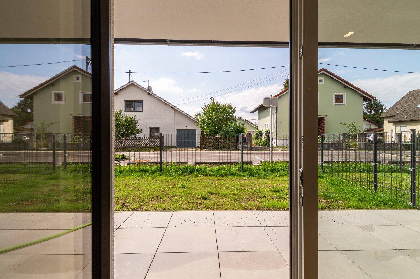 Blick von innen auf die gepflasterte Terrasse und den grünen Gartenbereich mit Nachbarhäusern.