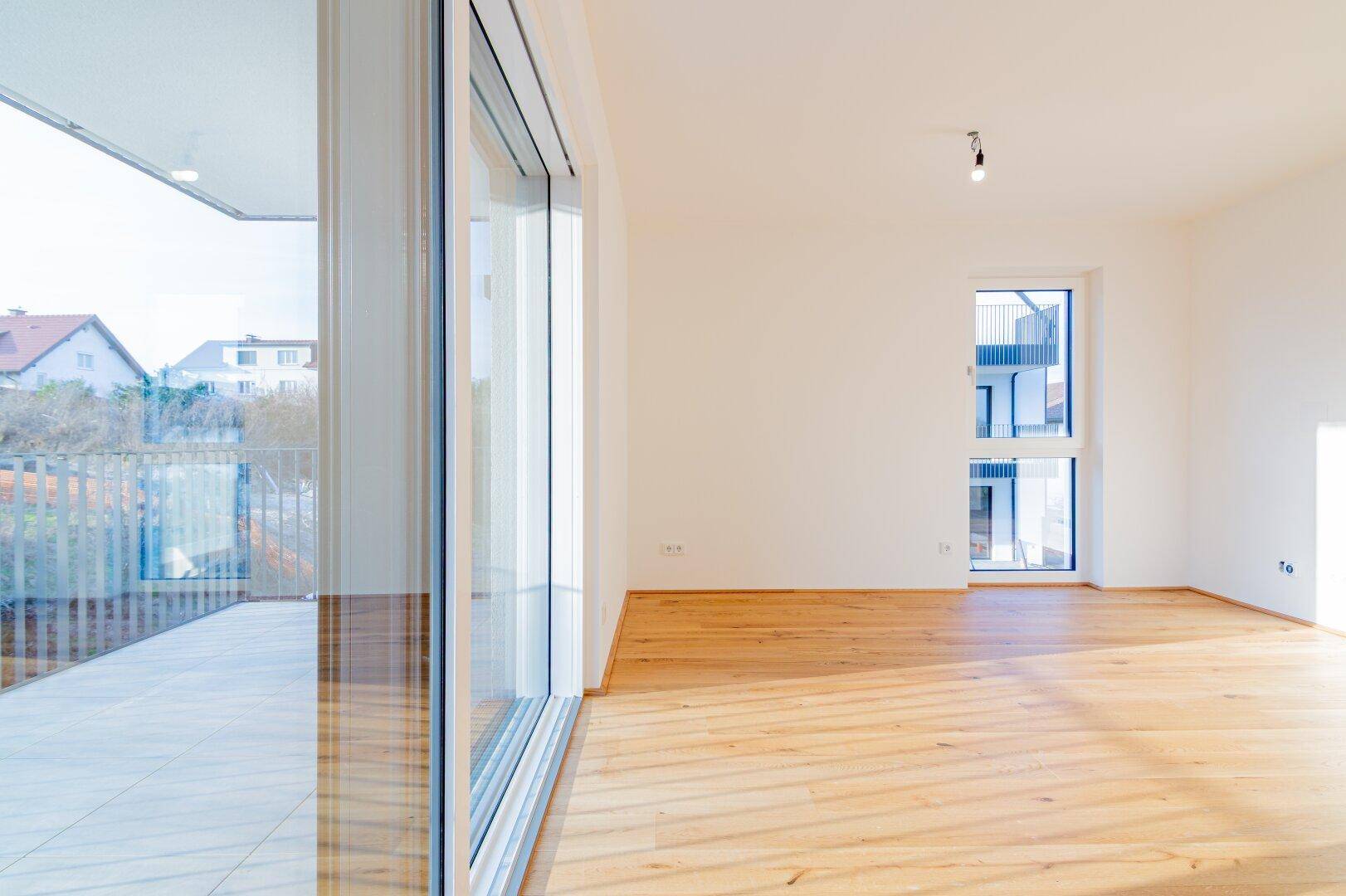 Geräumiger Raum mit Holzboden, großen Fenstern und Zugang zu einem Balkon oder einer Terrasse.