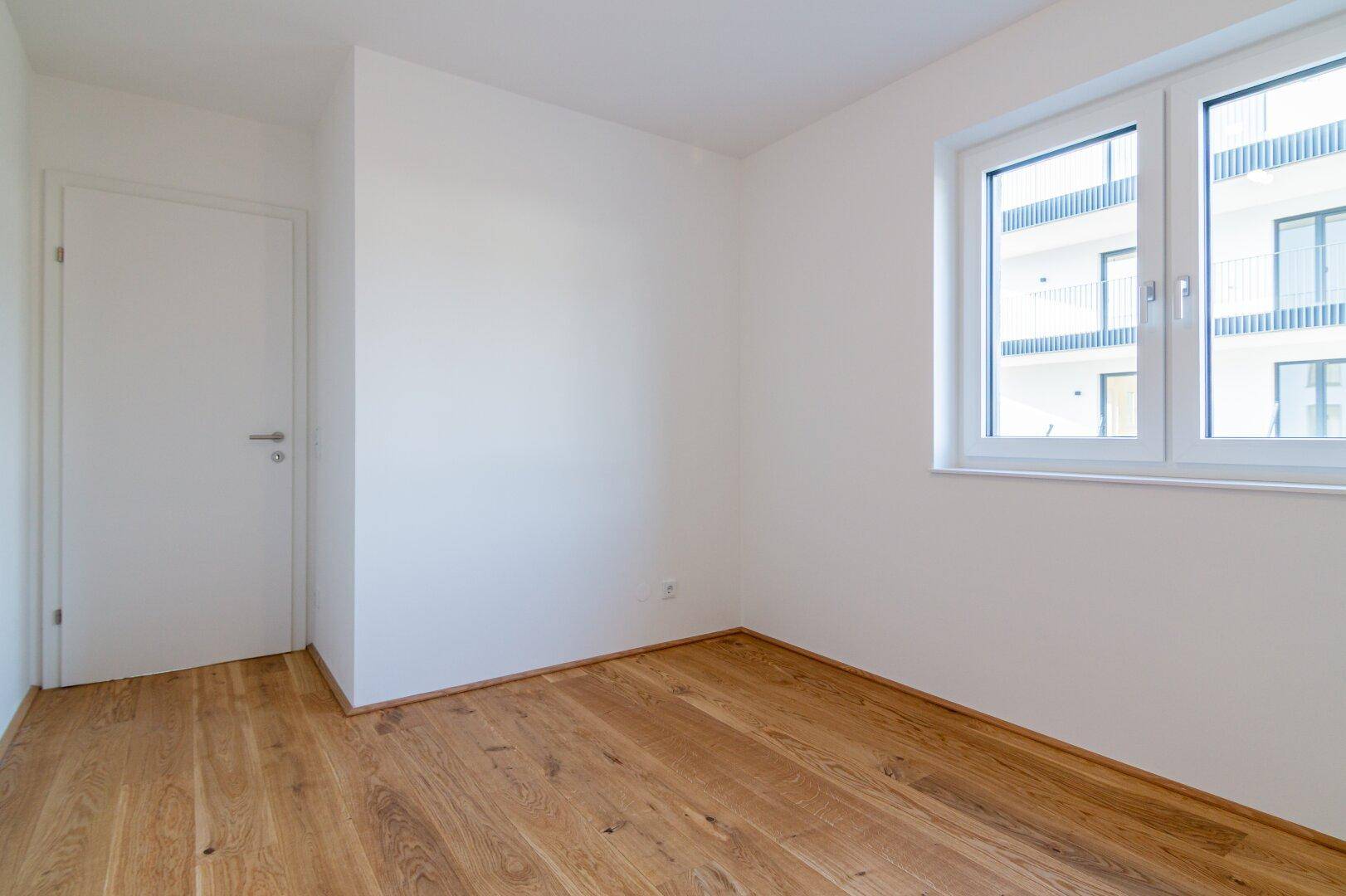 Freundliches Zimmer mit Holzboden und Fenster, das einen Blick auf ein gegenüberliegendes Gebäude bietet.