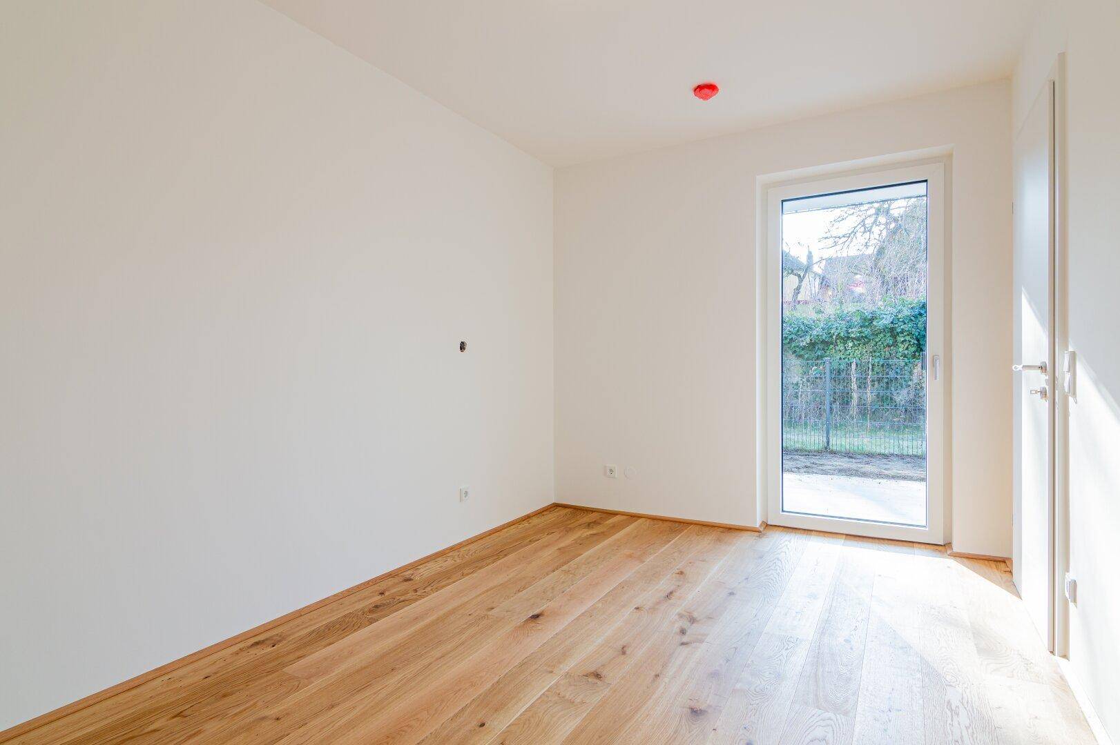 Sonniger Raum mit Holzboden und einer Glastür, die den Blick ins Grüne freigibt.