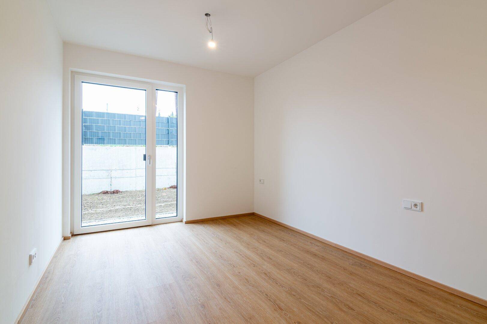 Leeres Schlafzimmer mit hellem Laminatboden und großem Fenster mit Zugang zum Außenbereich.