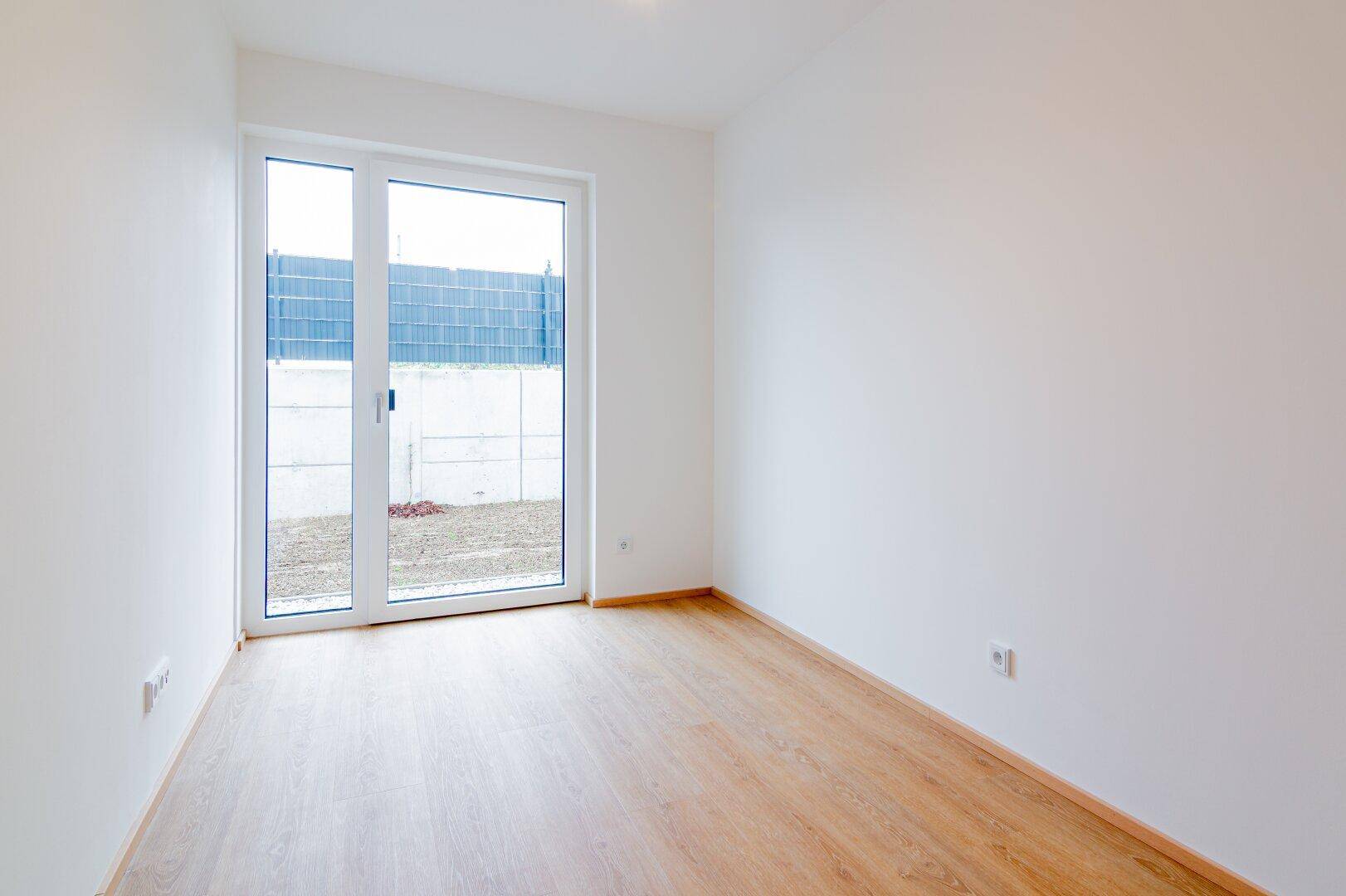 Leeres Zimmer mit hellem Laminatboden und großer Schiebetür, die zum Außenbereich führt.