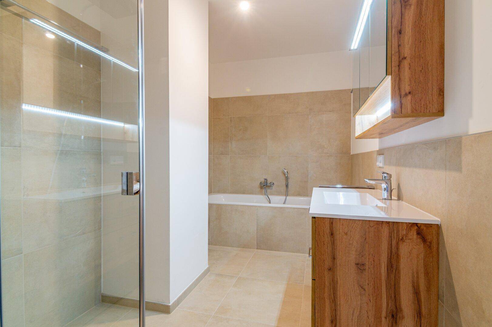 Modernes Badezimmer mit beigefarbenen Fliesen, Dusche, Badewanne und Holzwaschtisch.