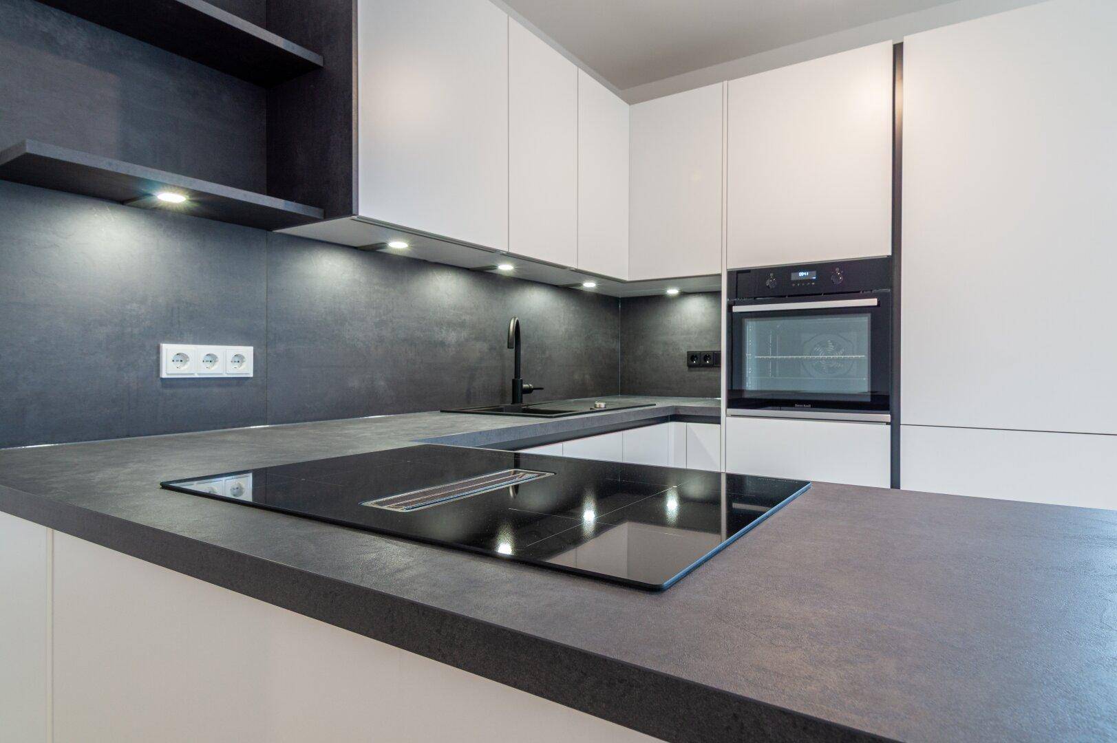 Moderne Einbauküche mit weißen Schränken, dunkler Arbeitsplatte und integriertem Induktionskochfeld.