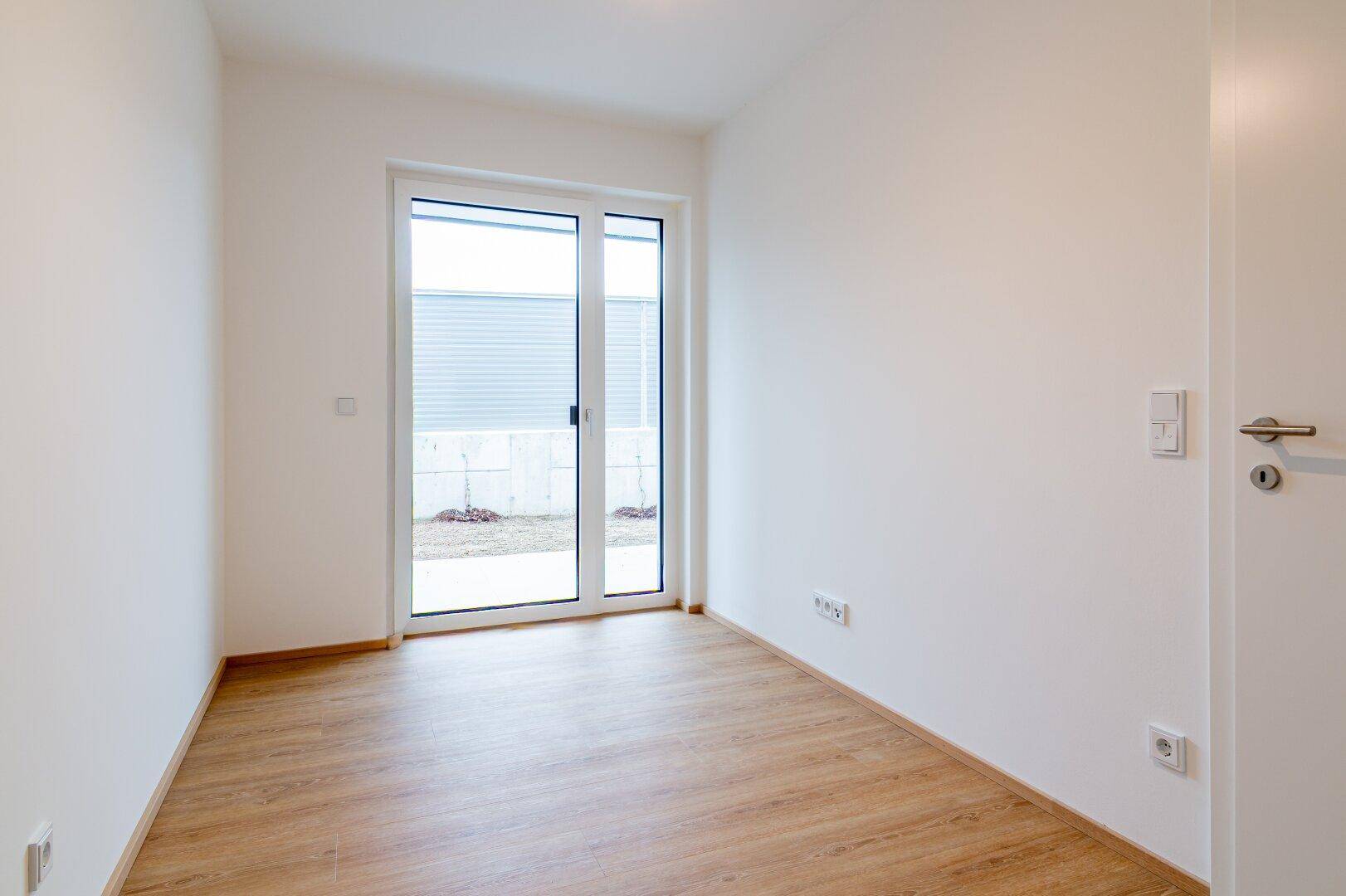 Leeres Zimmer mit hellem Laminatboden und großer Schiebetür, die zum Außenbereich führt.