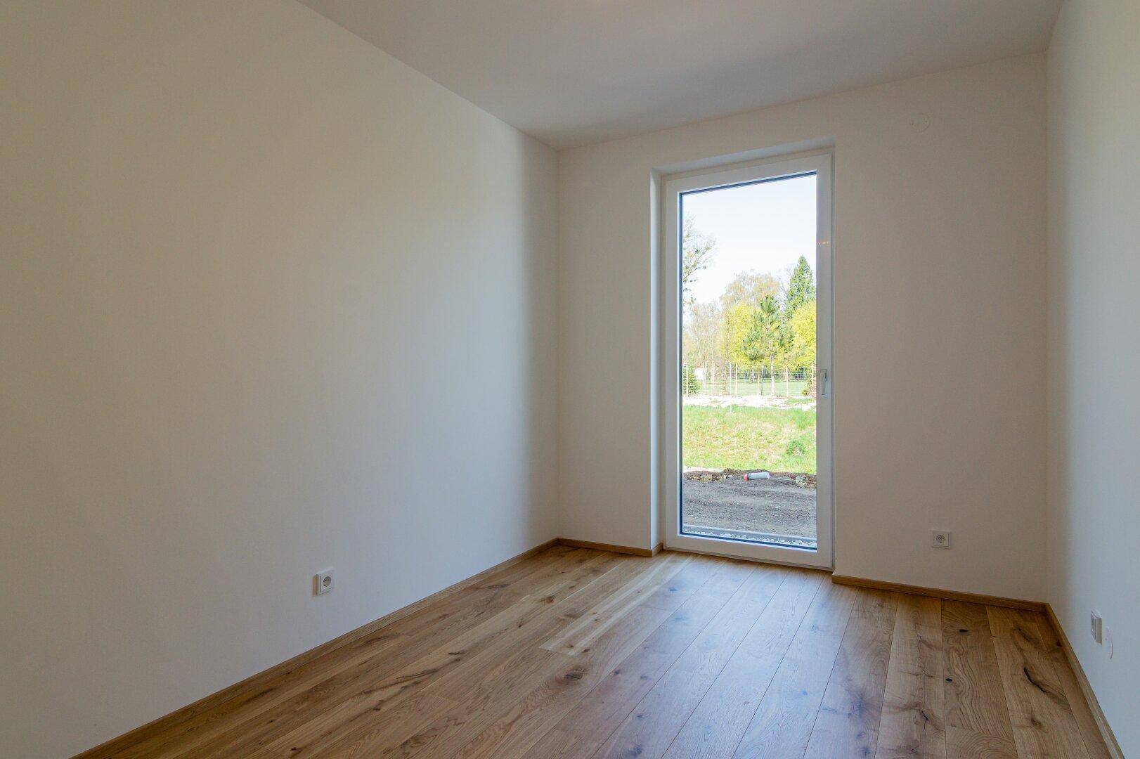 Leerer Raum mit Holzboden und großem Fenster mit Blick ins Grüne.