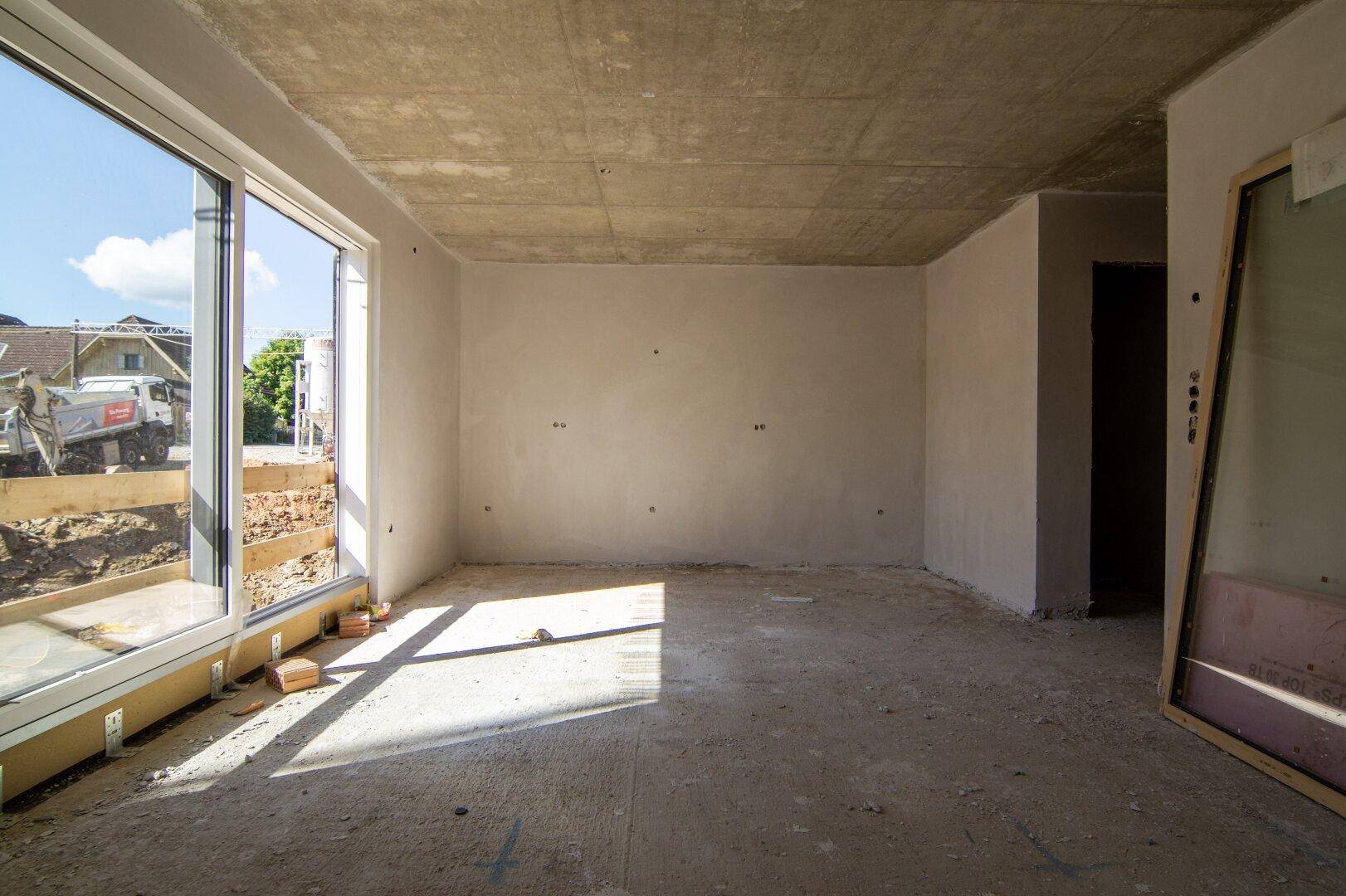 Unfertiger Innenraum mit großen Fenstern und Blick auf die Baustelle.