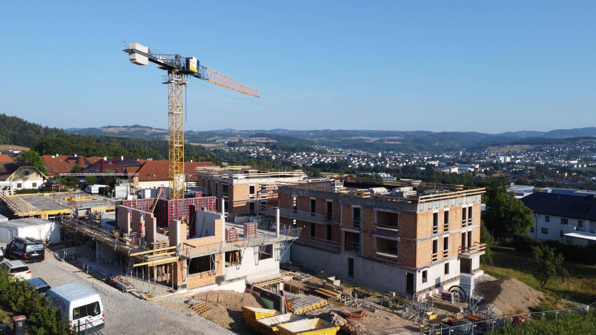 Übersicht einer großen Neubau-Baustelle mit mehreren Gebäuden und einem Baukran vor hügeliger Landschaft.