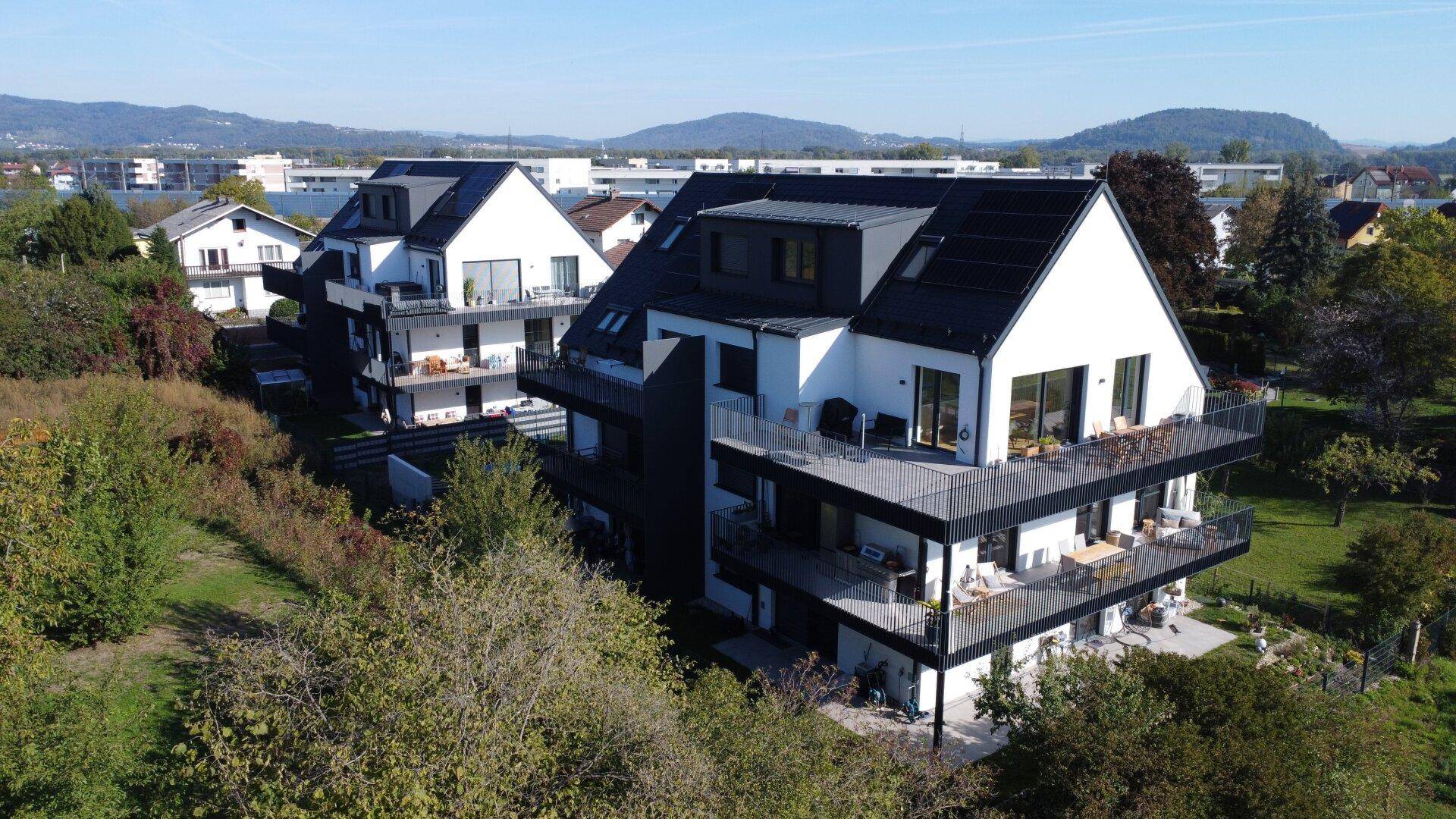 Moderne Wohngebäude mit Balkonen und Solaranlagen, eingebettet in grüne Landschaft mit Fernblick.