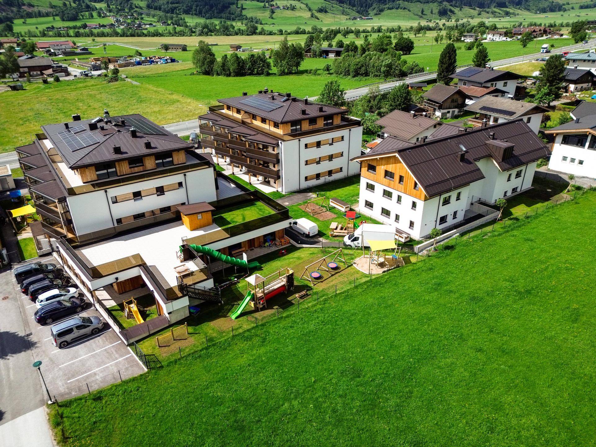 Luftaufnahme von Neubauten mit Spielplatz und Parkplätzen, umgeben von grünen Wiesen und Bergen.