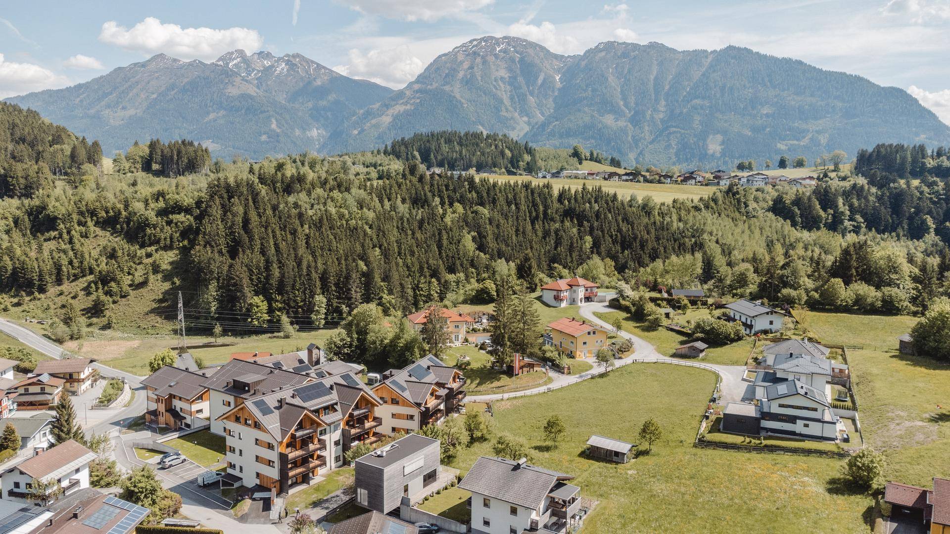 Weitläufige Luftaufnahme einer ländlichen Siedlung, eingebettet in grüne Natur mit Wäldern und majestätischen Bergen im Hintergrund.