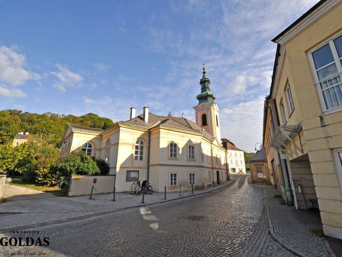 Historische Straße mit Kopfsteinpflaster, einem gelben Altbau und einem Kirchturm unter blauem Himmel.