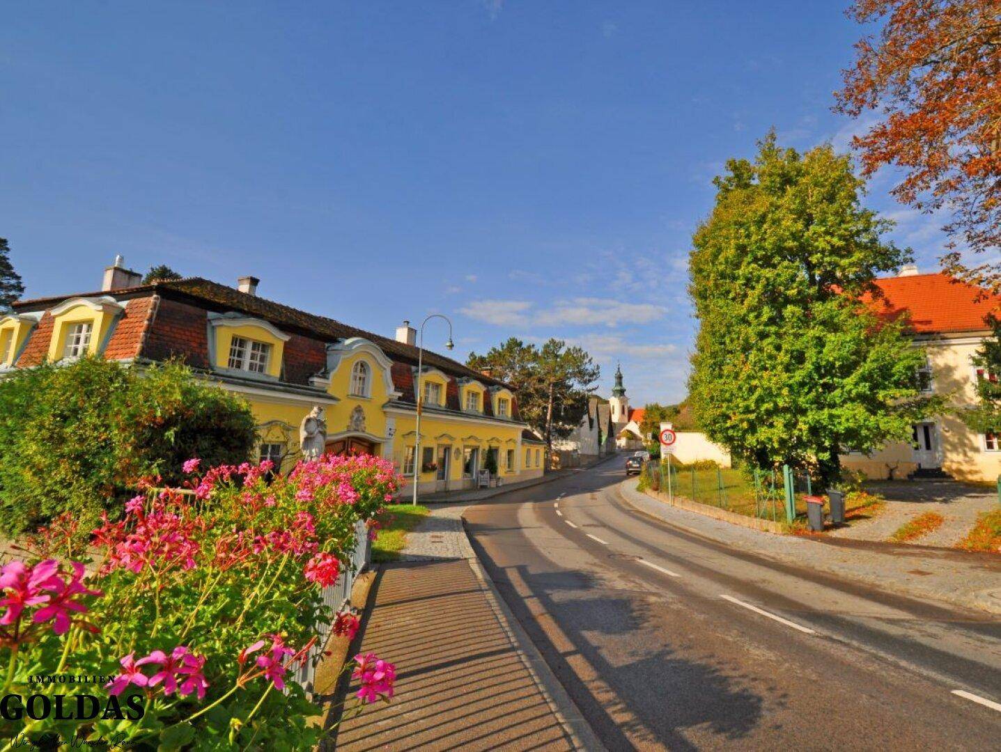 Belebte Straße mit historischen Gebäuden, bunten Blumen und einem Kirchturm in der Ferne.