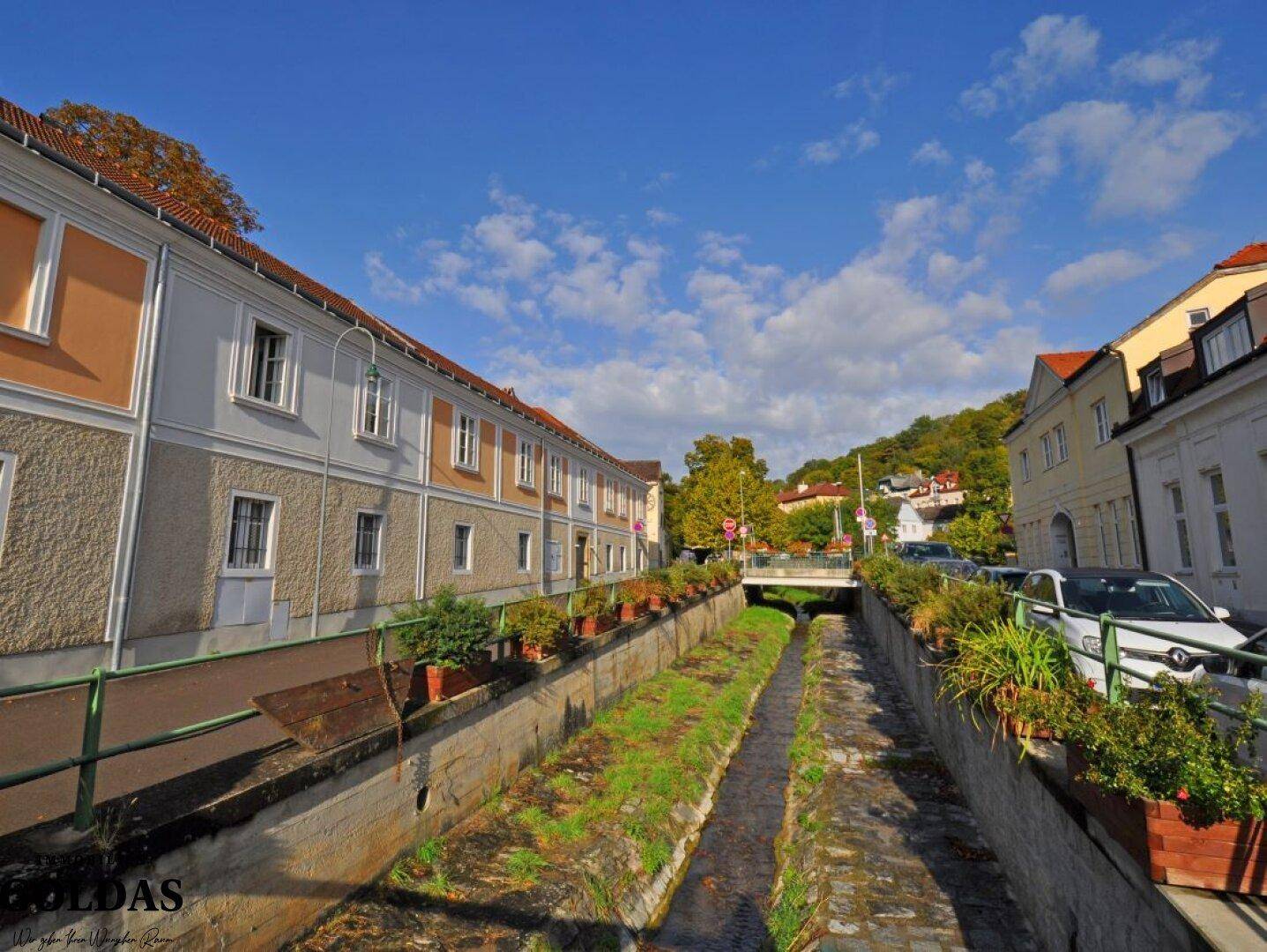 Straße entlang eines Kanals mit historischen Gebäuden und geparkten Autos unter blauem Himmel.