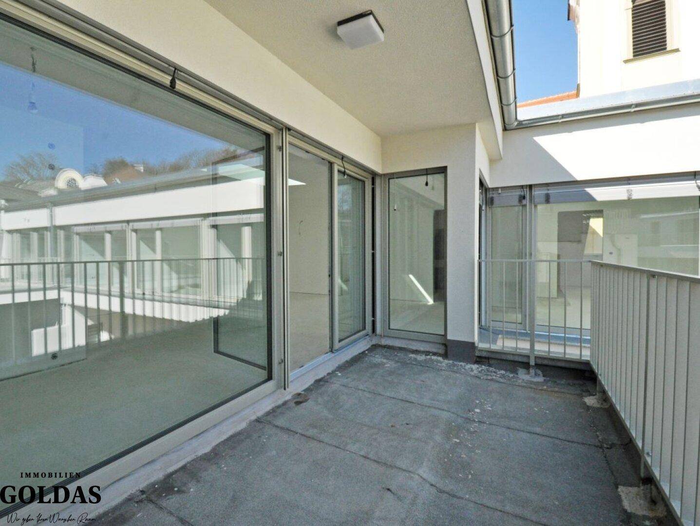 Geräumiger Balkon mit Betonboden und Glas-Schiebetüren zum Innenbereich des Neubaus.
