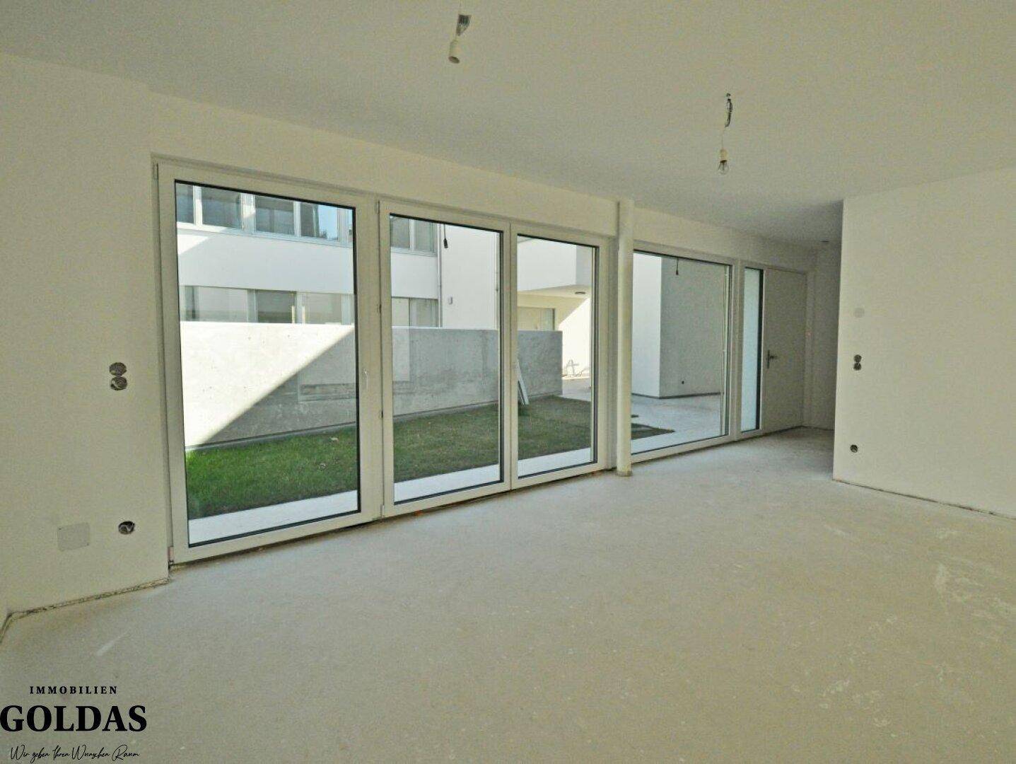 Heller Innenraum im Rohbauzustand mit großen Fensterfronten und Blick auf die private Grünfläche.