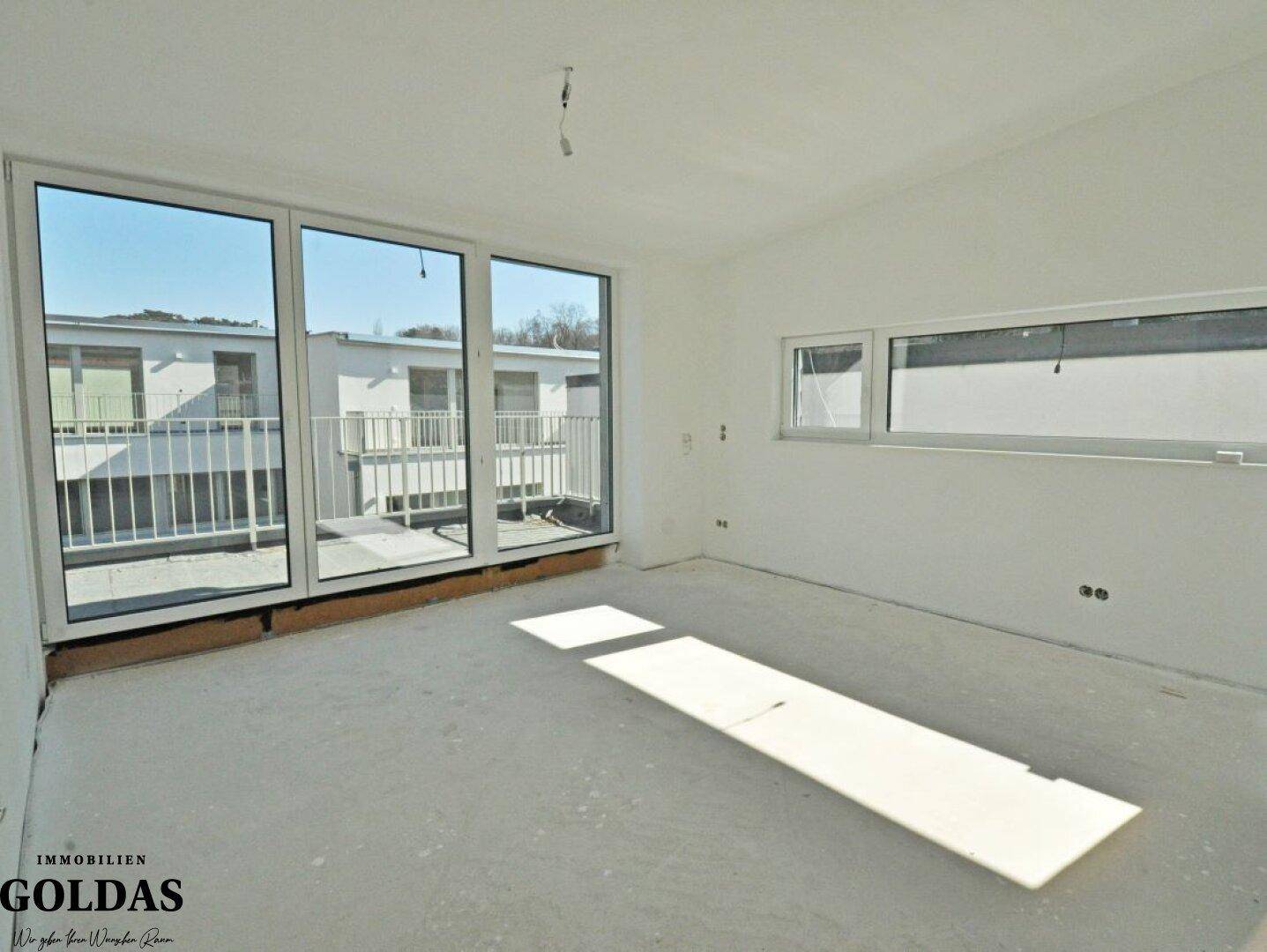 Heller Raum im Rohbau mit großen Fenstern und Zugang zu einem Außenbereich, der viel Tageslicht bietet.