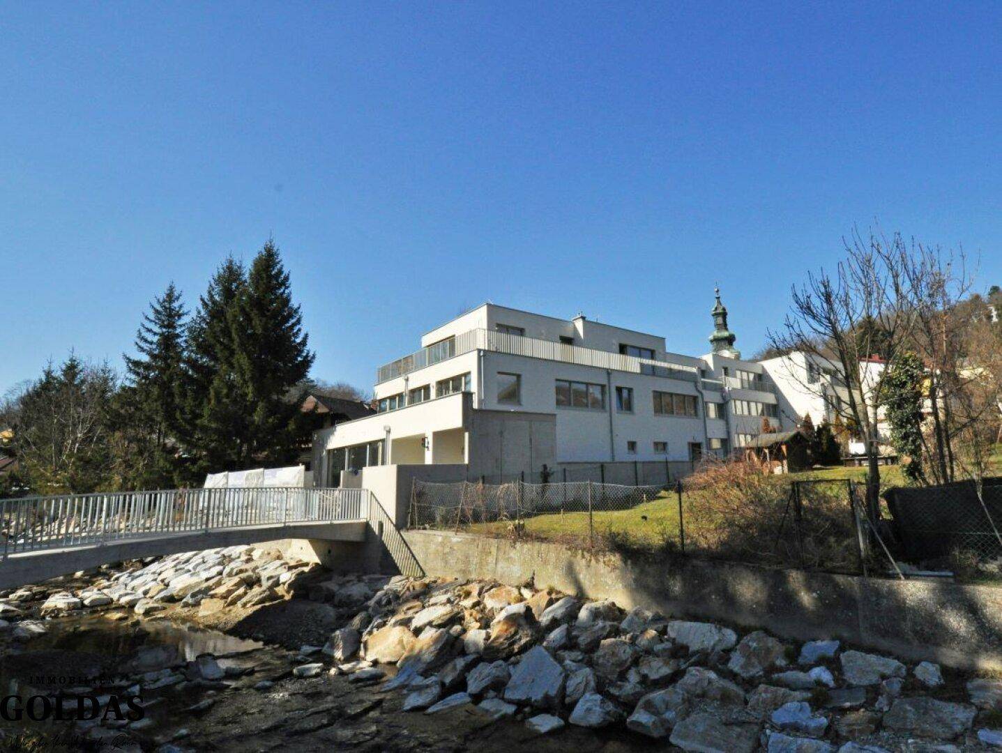 Moderne Wohnanlage am Flussufer mit Brücke und Blick auf die umliegende Natur und einen Kirchturm.