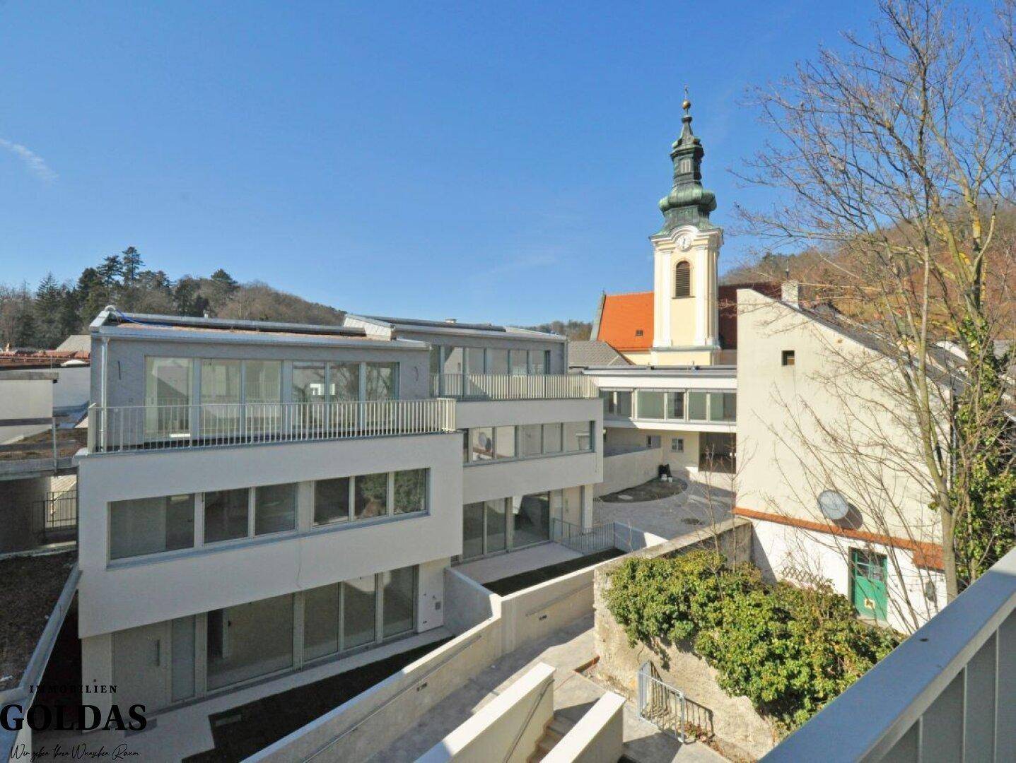 Gesamtansicht des modernen Wohnkomplexes mit angrenzender Kirche und hügeliger Landschaft im Hintergrund.