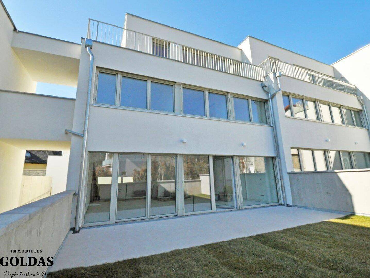 Moderne Gebäudefassade mit großen Glasfronten, angrenzender Terrasse und gepflegtem Rasenbereich.