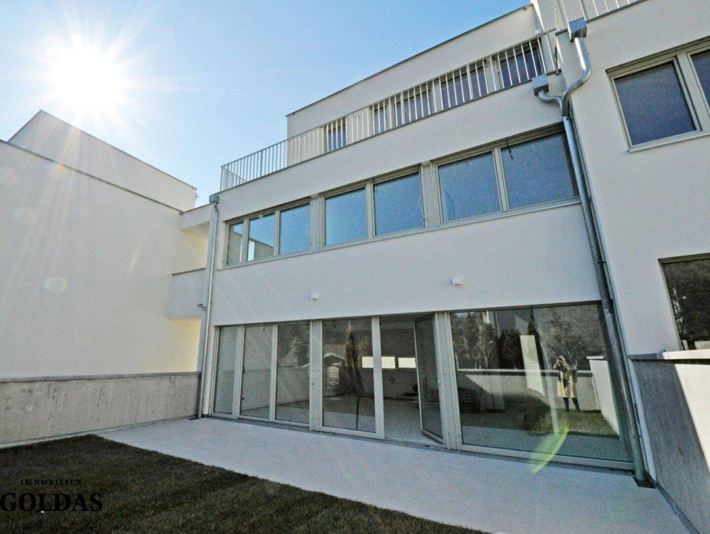 Sonnige Ansicht der modernen Gebäudefassade mit großen Fenstern, Terrasse und grünem Rasen.