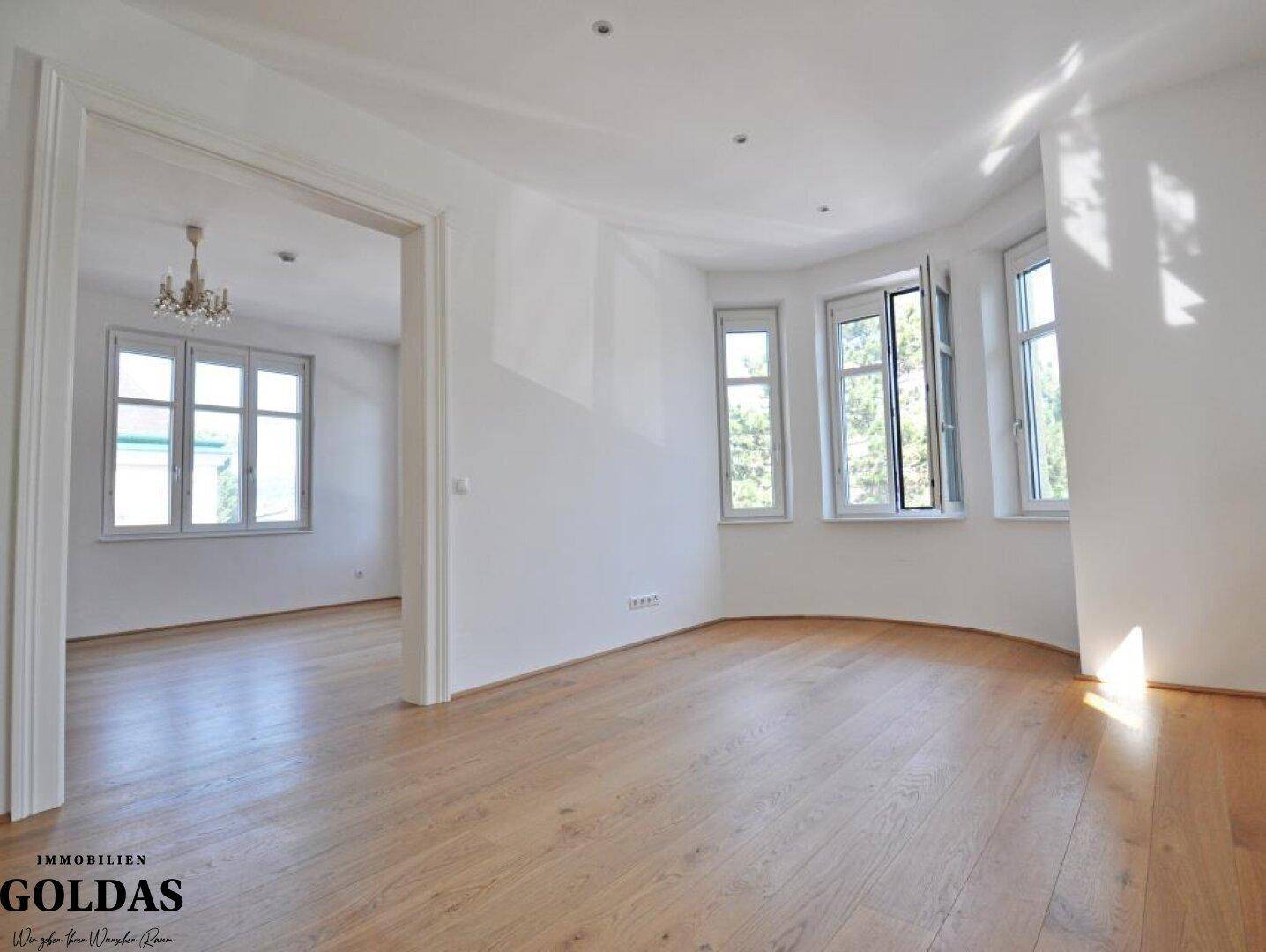 Helles Wohnzimmer mit Holzboden, großen Fenstern und Zugang zu einem weiteren Raum.