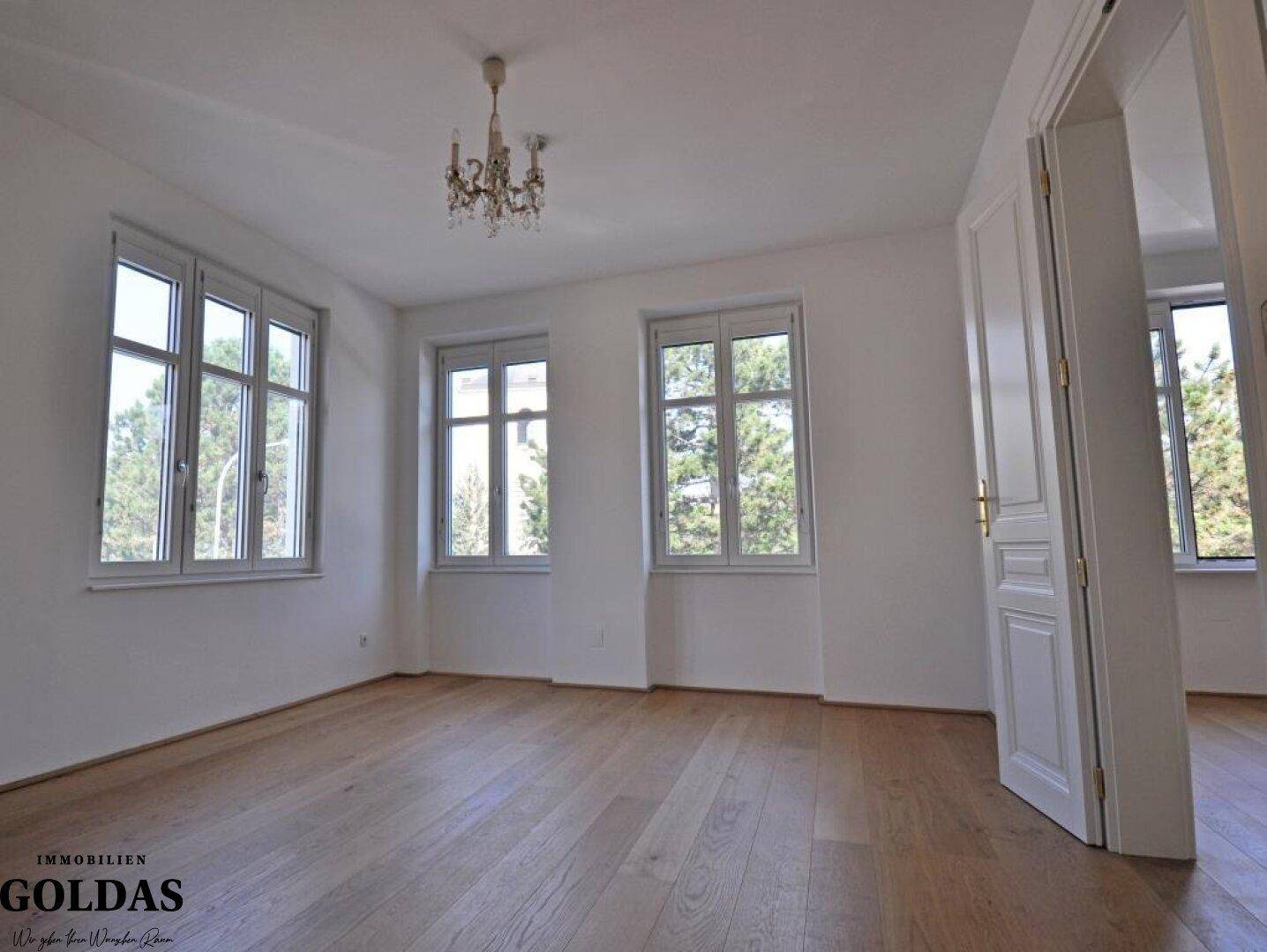 Geräumiges Wohnzimmer mit Holzboden, Kronleuchter und mehreren Fenstern mit Grünblick.