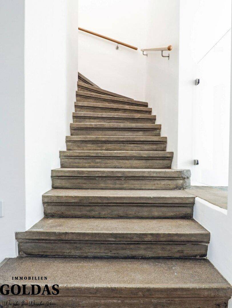 Elegante Steintreppe mit schlichtem Holzgeländer in einem hellen Treppenhaus.