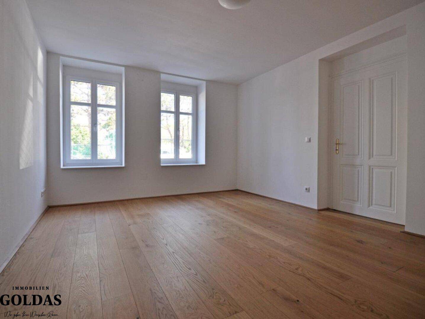 Helles, leeres Zimmer mit Holzboden und zwei Fenstern, bereit zur individuellen Gestaltung.