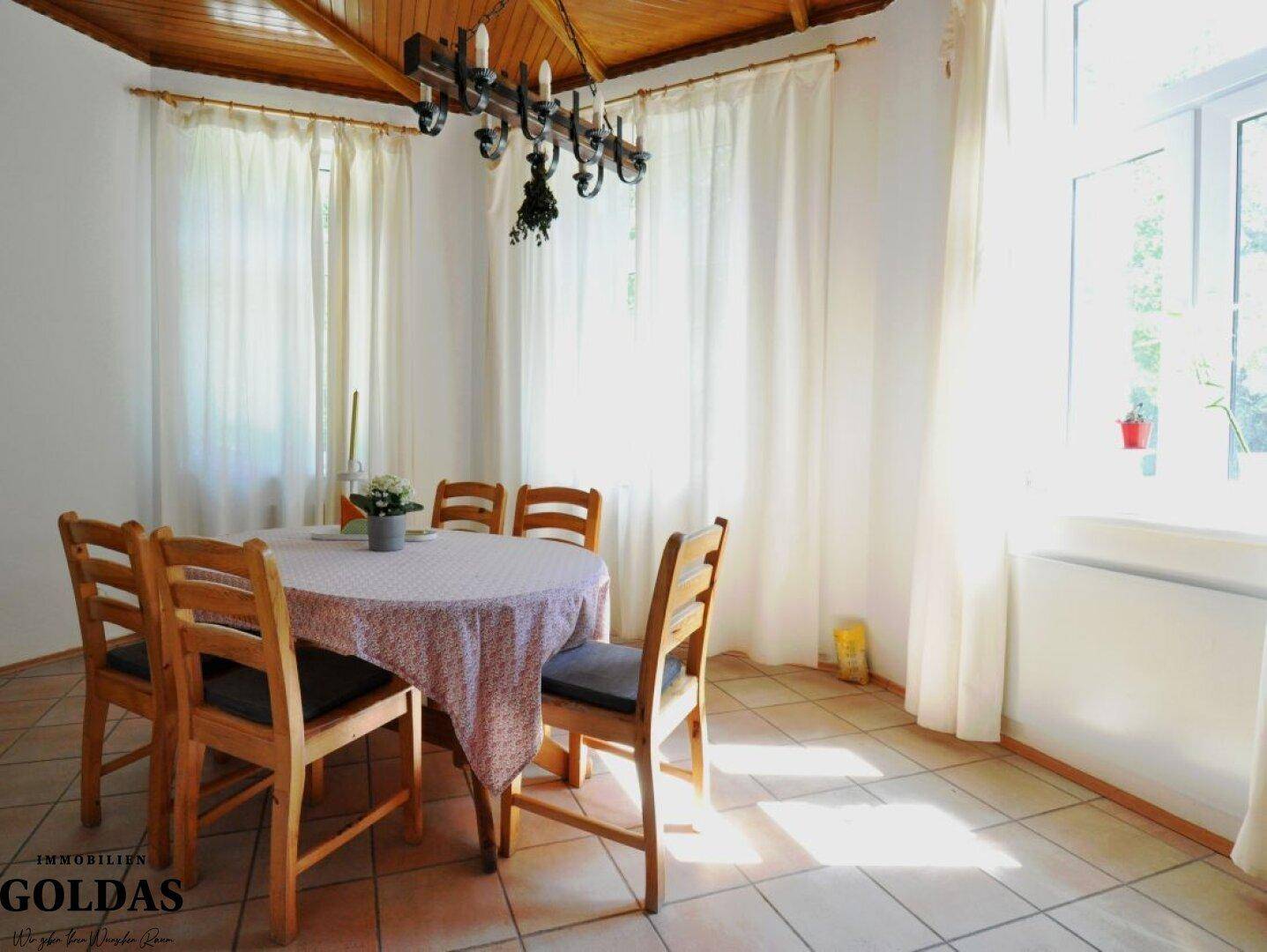 Heller Essbereich mit Holztisch, Stühlen und Fliesenboden unter einer Holzbalkendecke.
